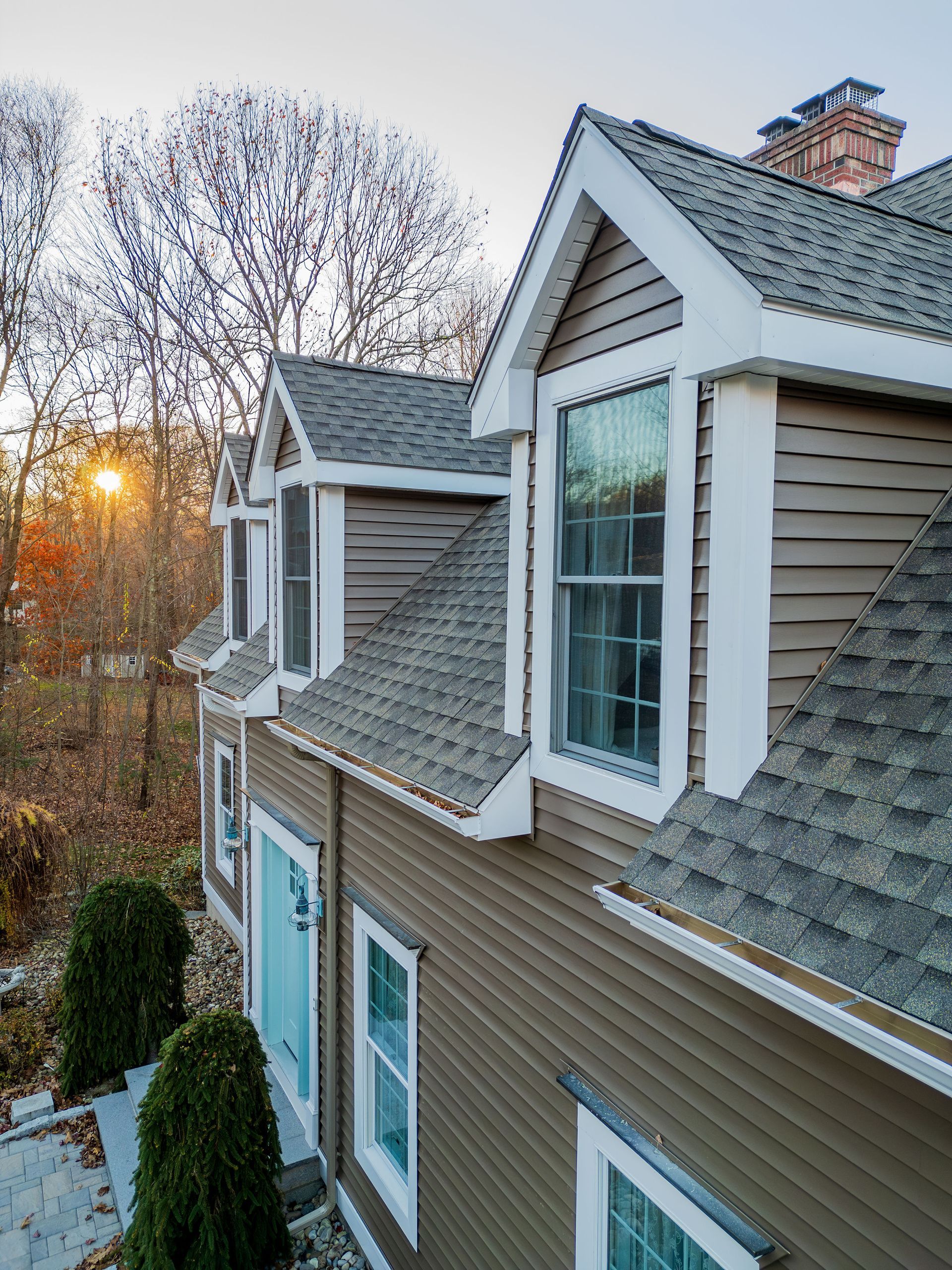 Brown house with white trim, multiple windows, and a chimney against a backdrop of trees and sunset.