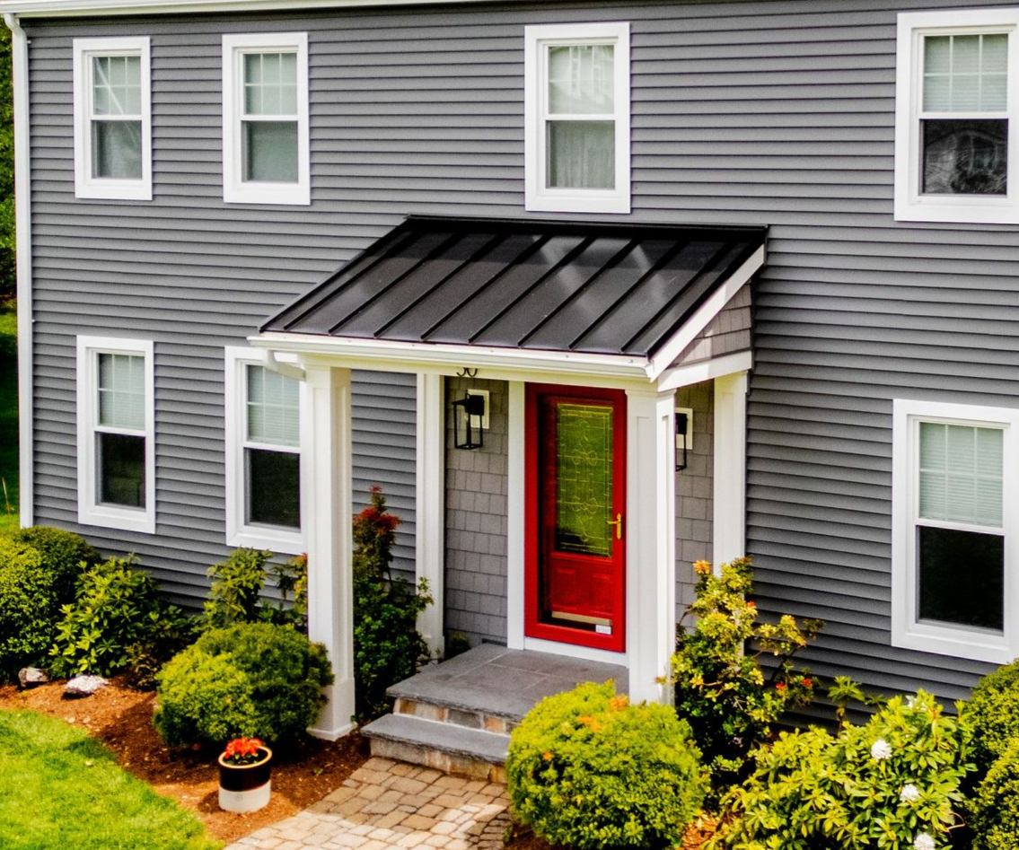 Gray house with a red door and a black awning, surrounded by greenery.