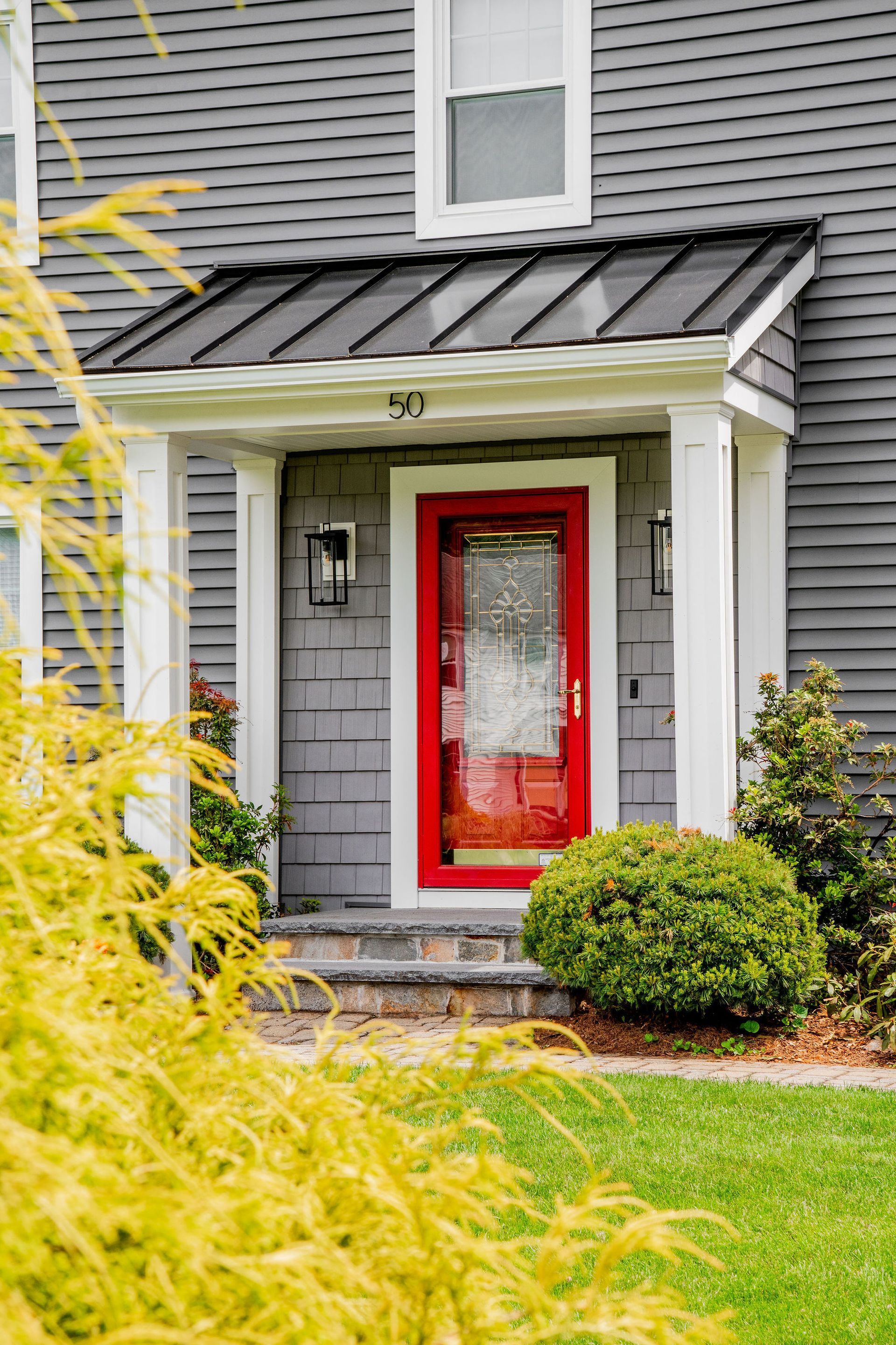 Front porch with bright red door, gray siding, and trimmed shrubs in front of a house