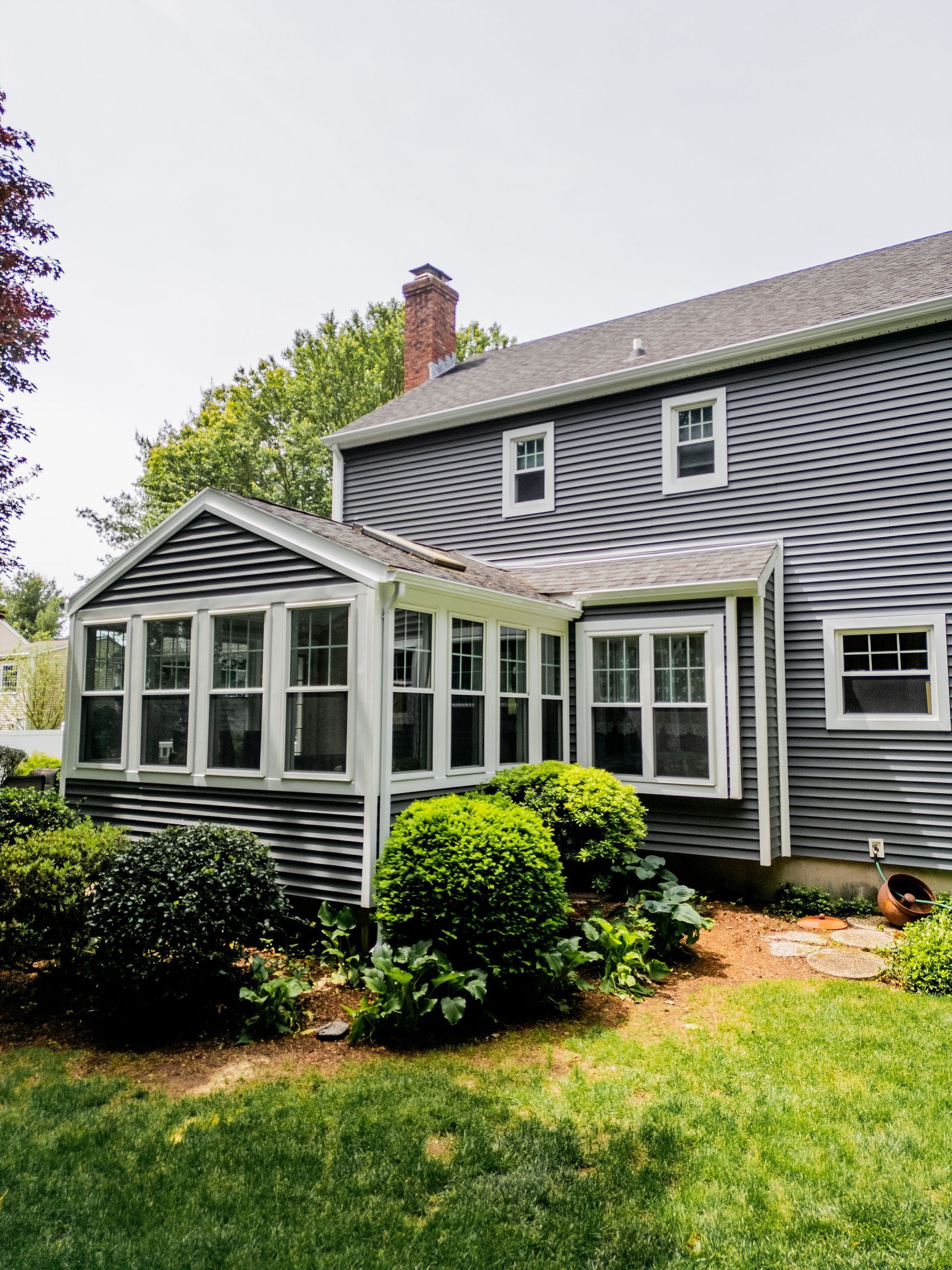 Side view of a gray shingled house with a sunroom, surrounded by green bushes and grass.