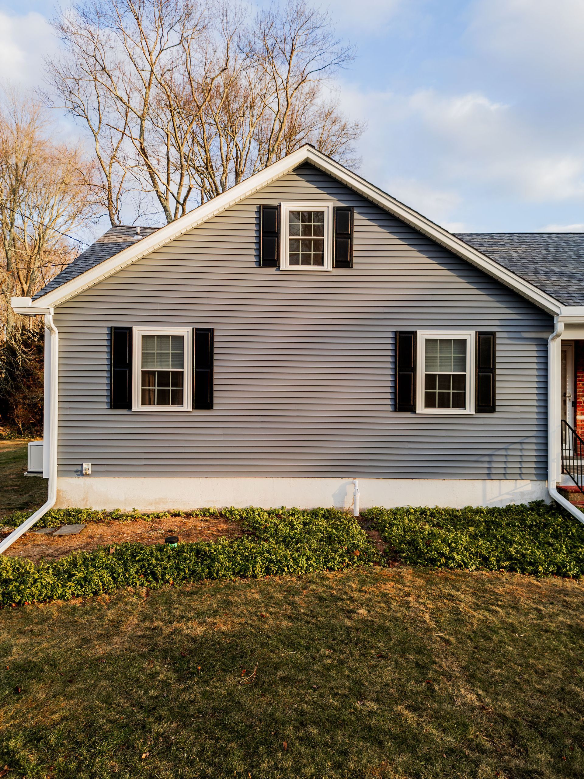 Gray house with black shutters, two windows on the lower level, one above. Brown grass and green plants in front.