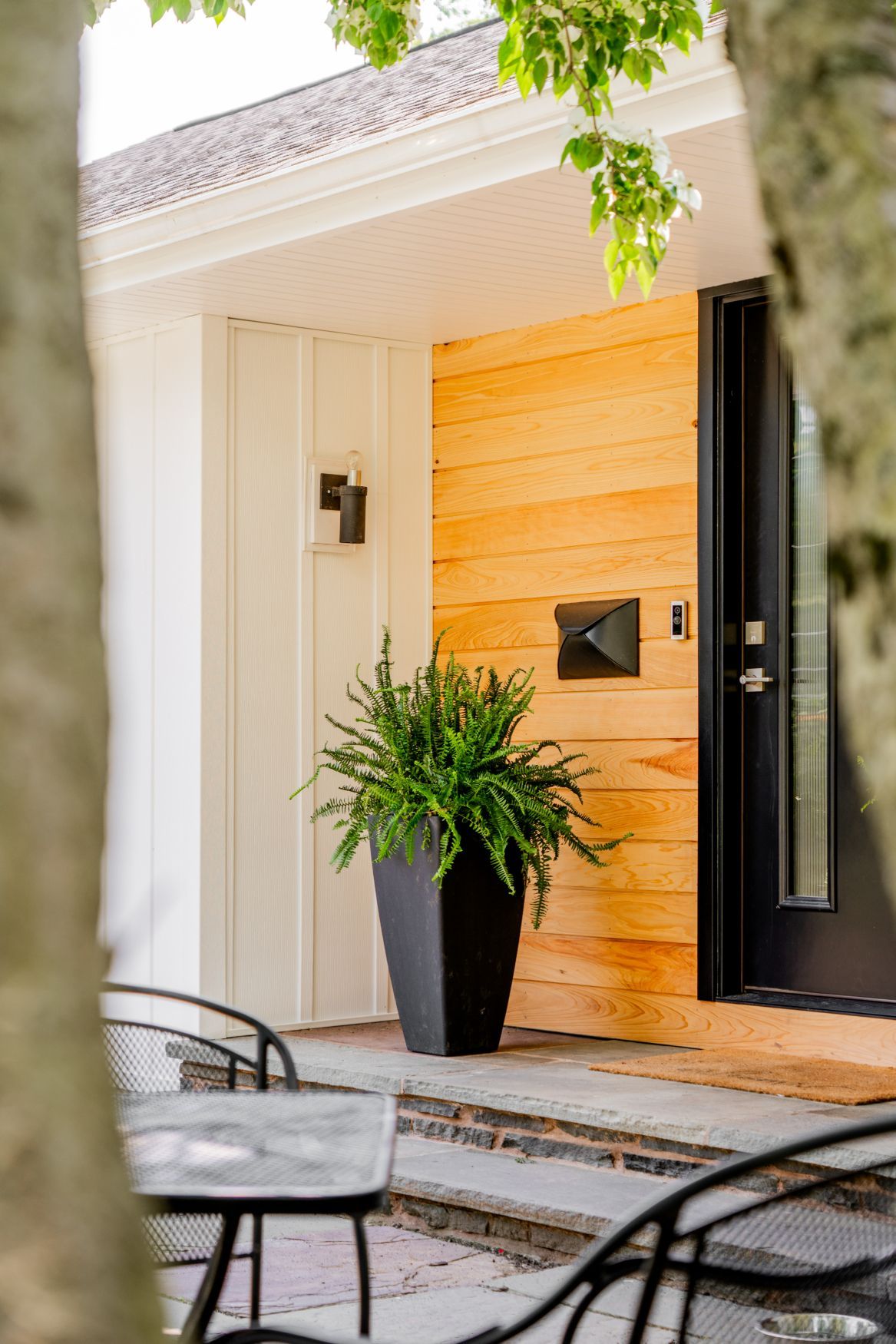 Modern front porch with black door, wood siding, potted plant, and metal patio chairs