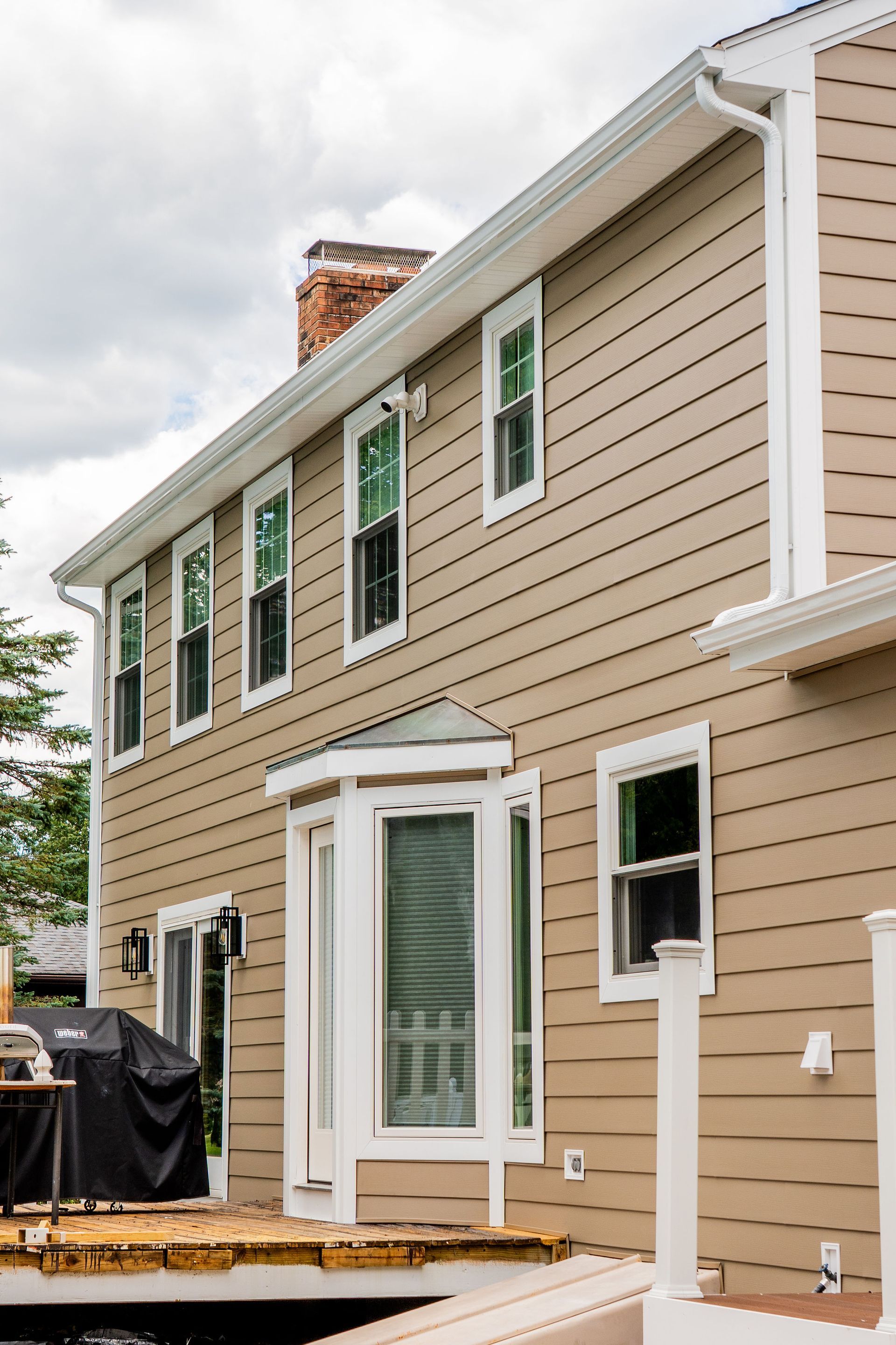 Tan house with white trim, windows, deck, and grill. Cloudy sky.