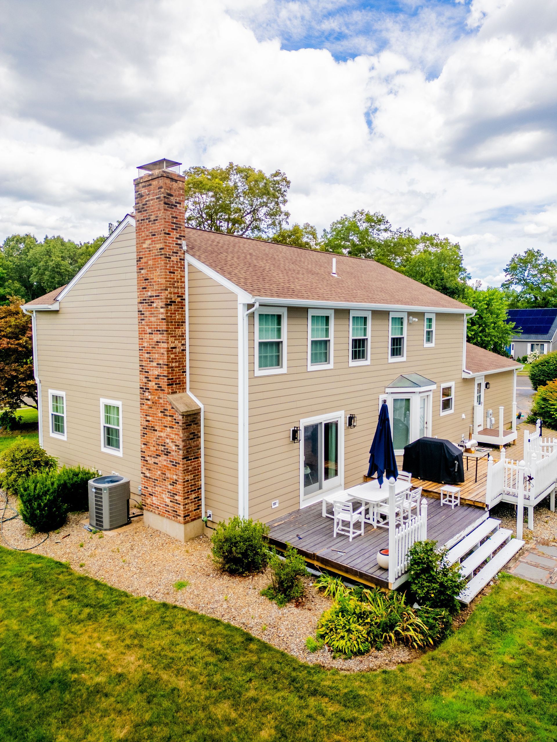 Two-story beige house with brick chimney and deck, surrounded by green lawn and shrubs.