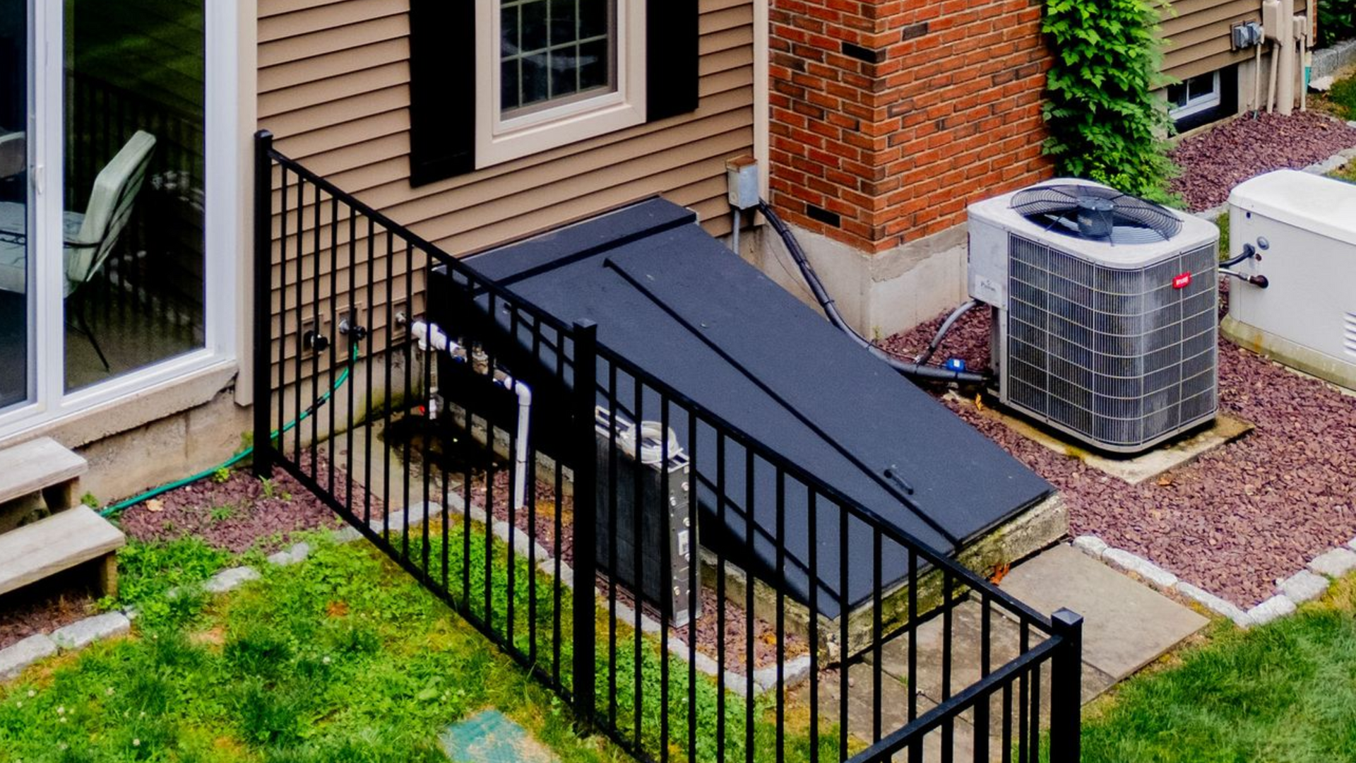 Black metal fence surrounds a dark cover over a basement entrance, next to an air conditioning unit and brick chimney.