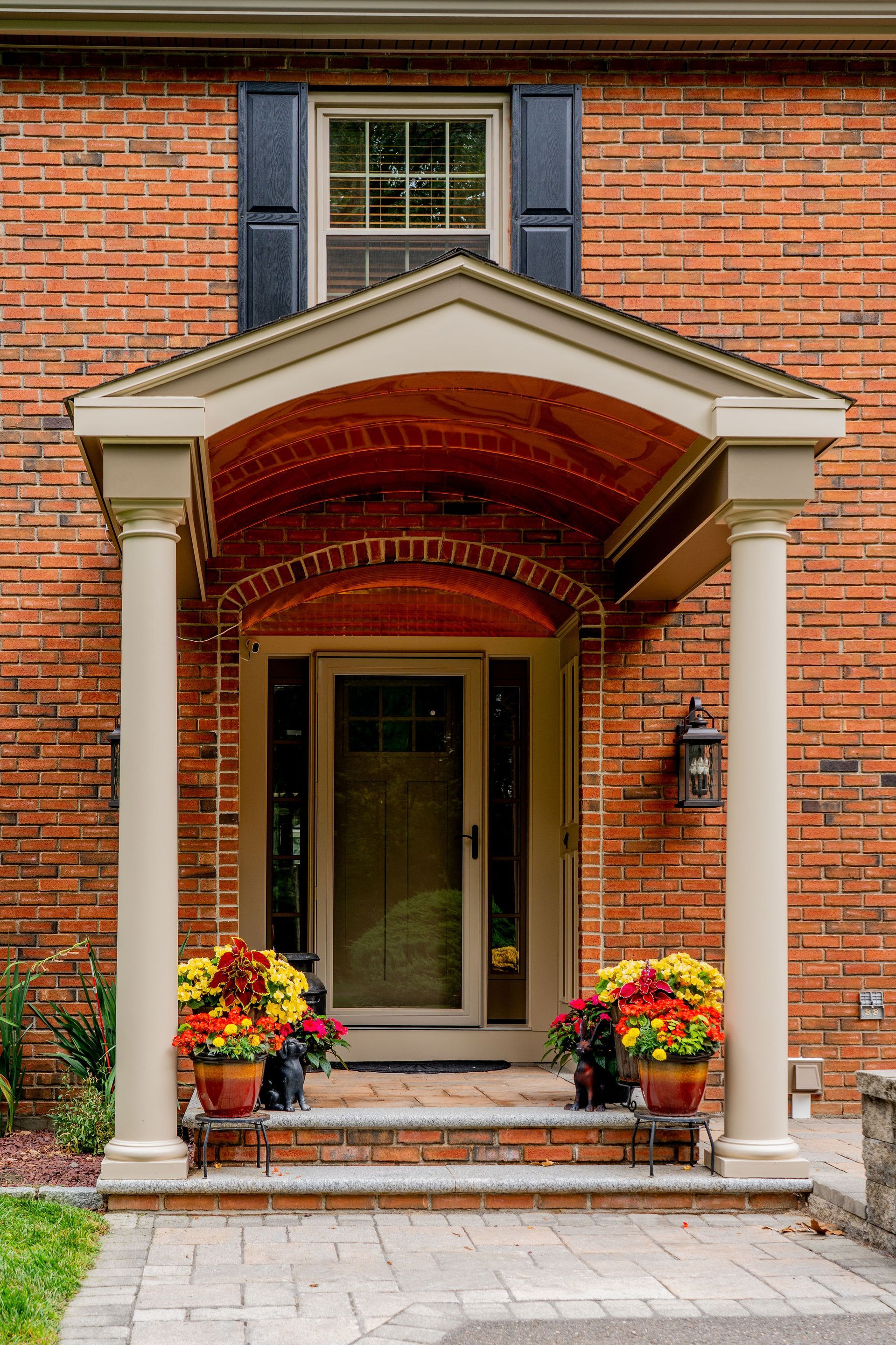 Red brick house entrance with beige columns, arched roof, glass door, and potted mums.