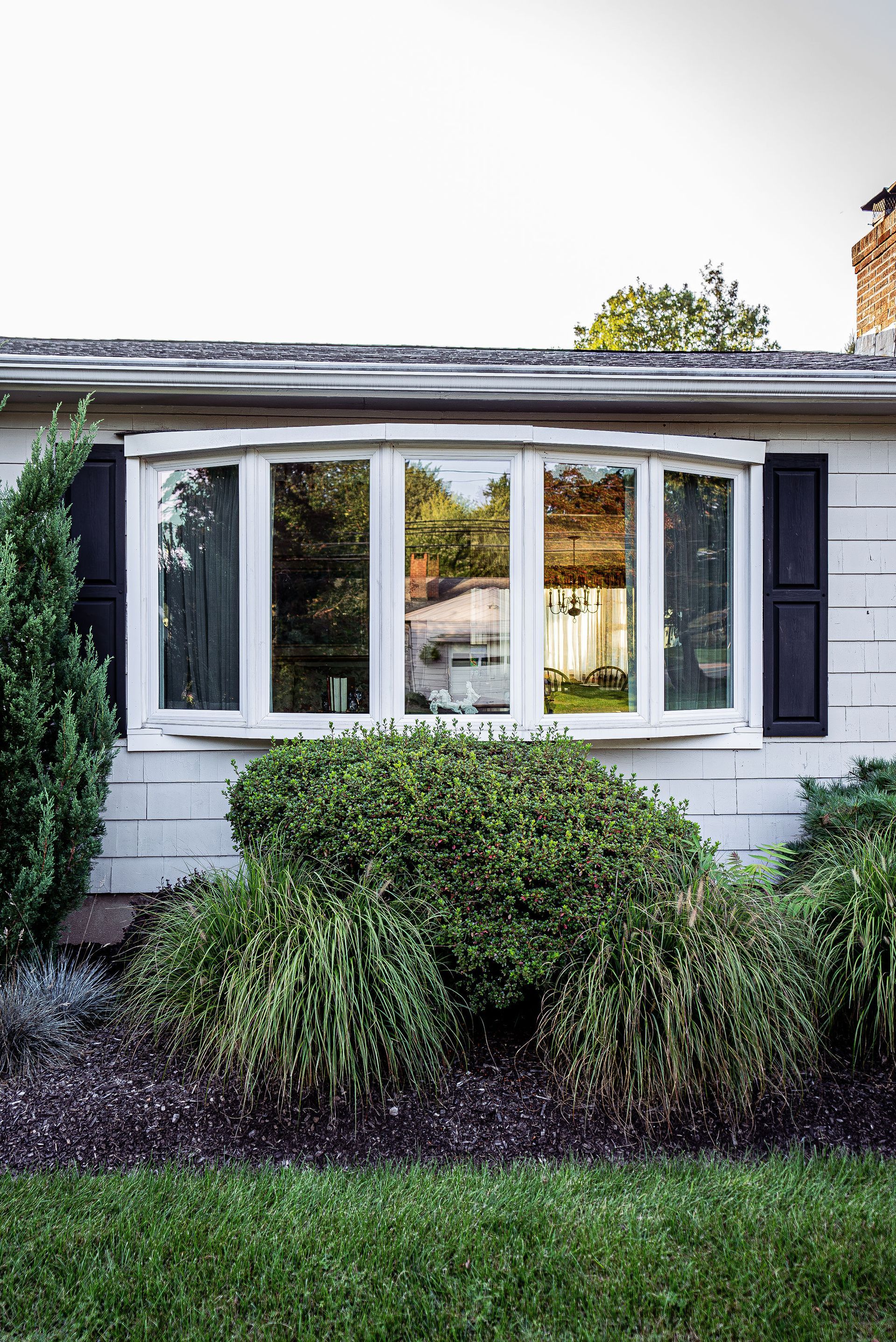 White house exterior with bay window, black shutters, and overgrown bushes.