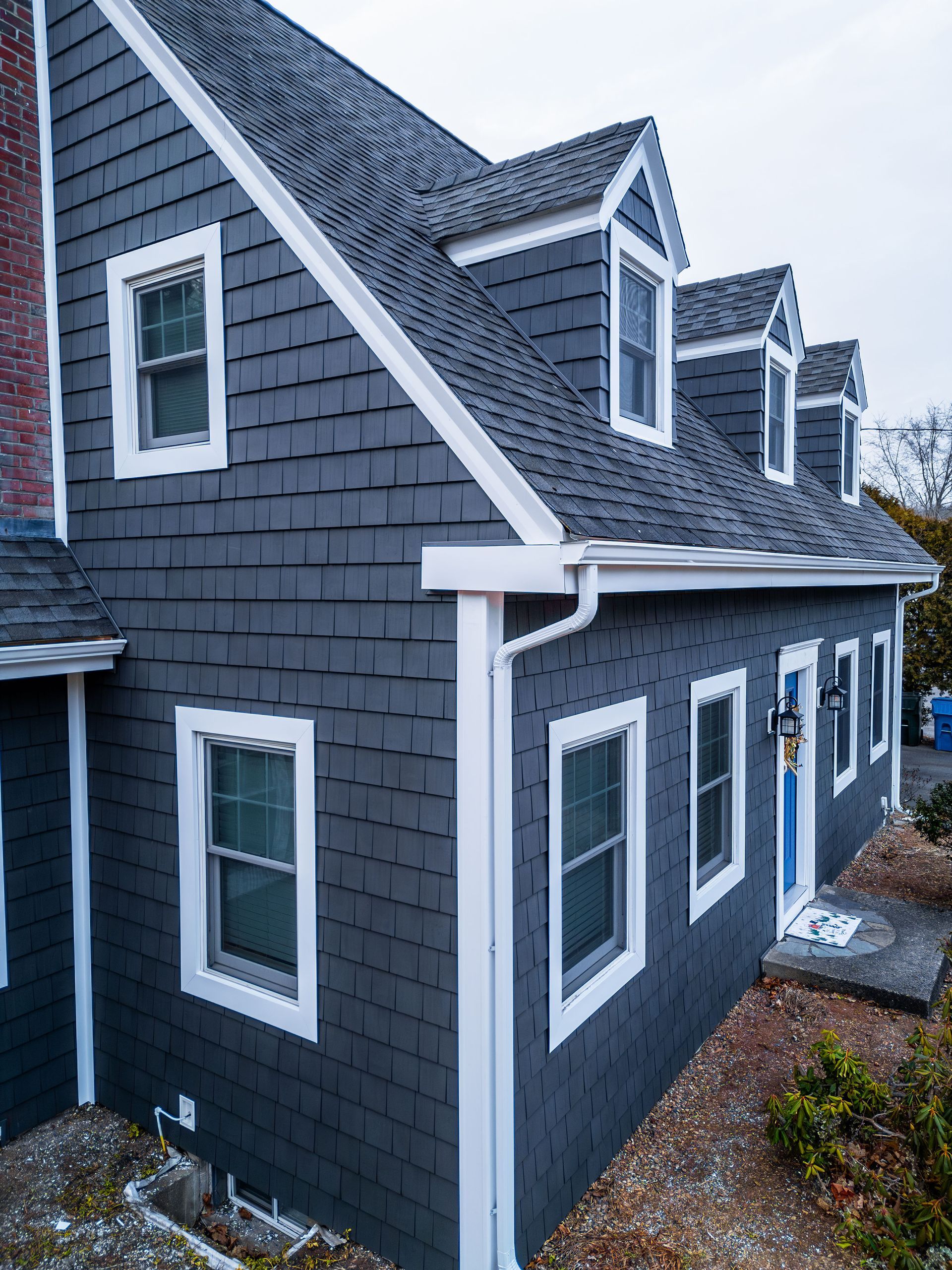 Gray shingle siding house with white trim, dormers, and windows. Brick chimney visible.
