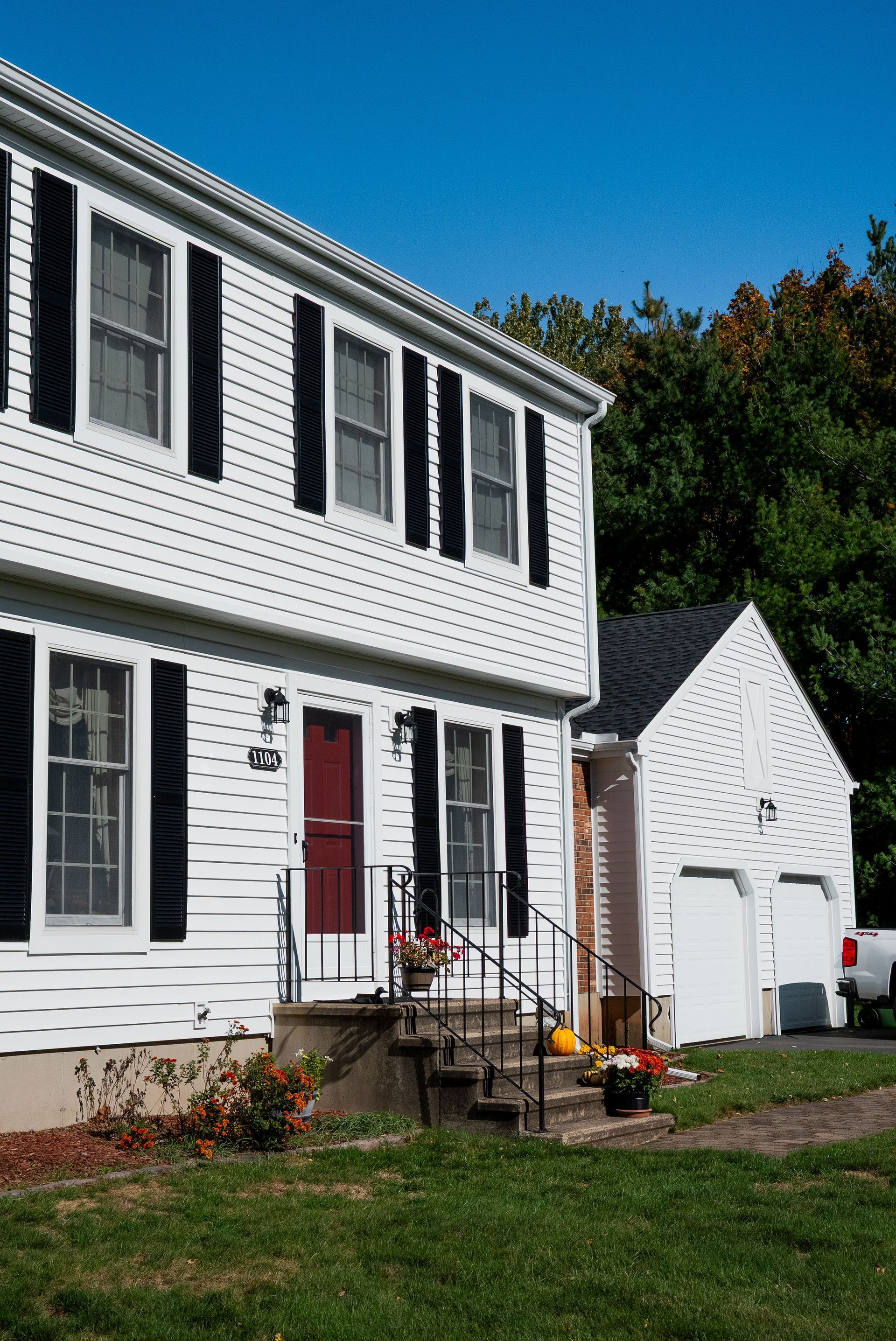 A white two-story house with black shutters, a red front door, and an attached two-car garage under a clear blue sky.