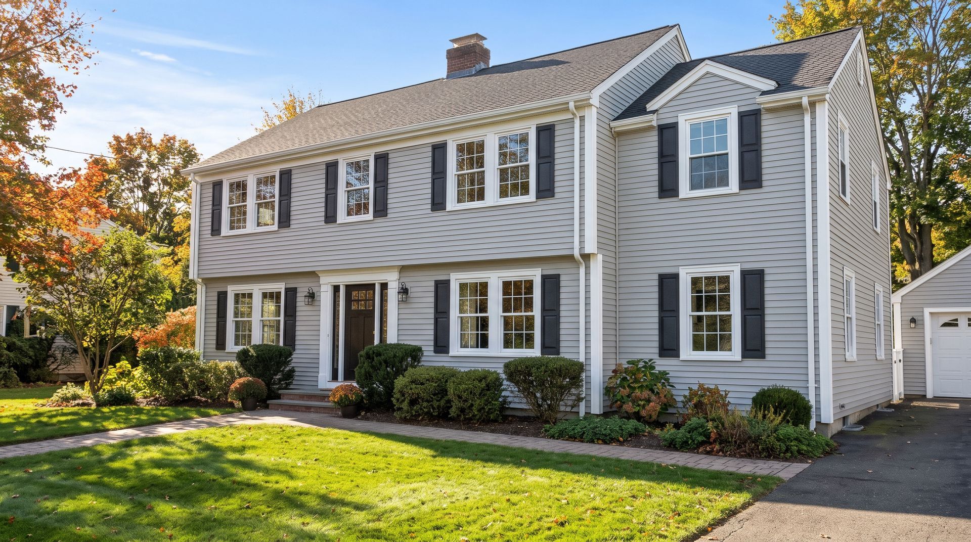 White two-story suburban house with black shutters, a front lawn, and a driveway in autumn sunlight
