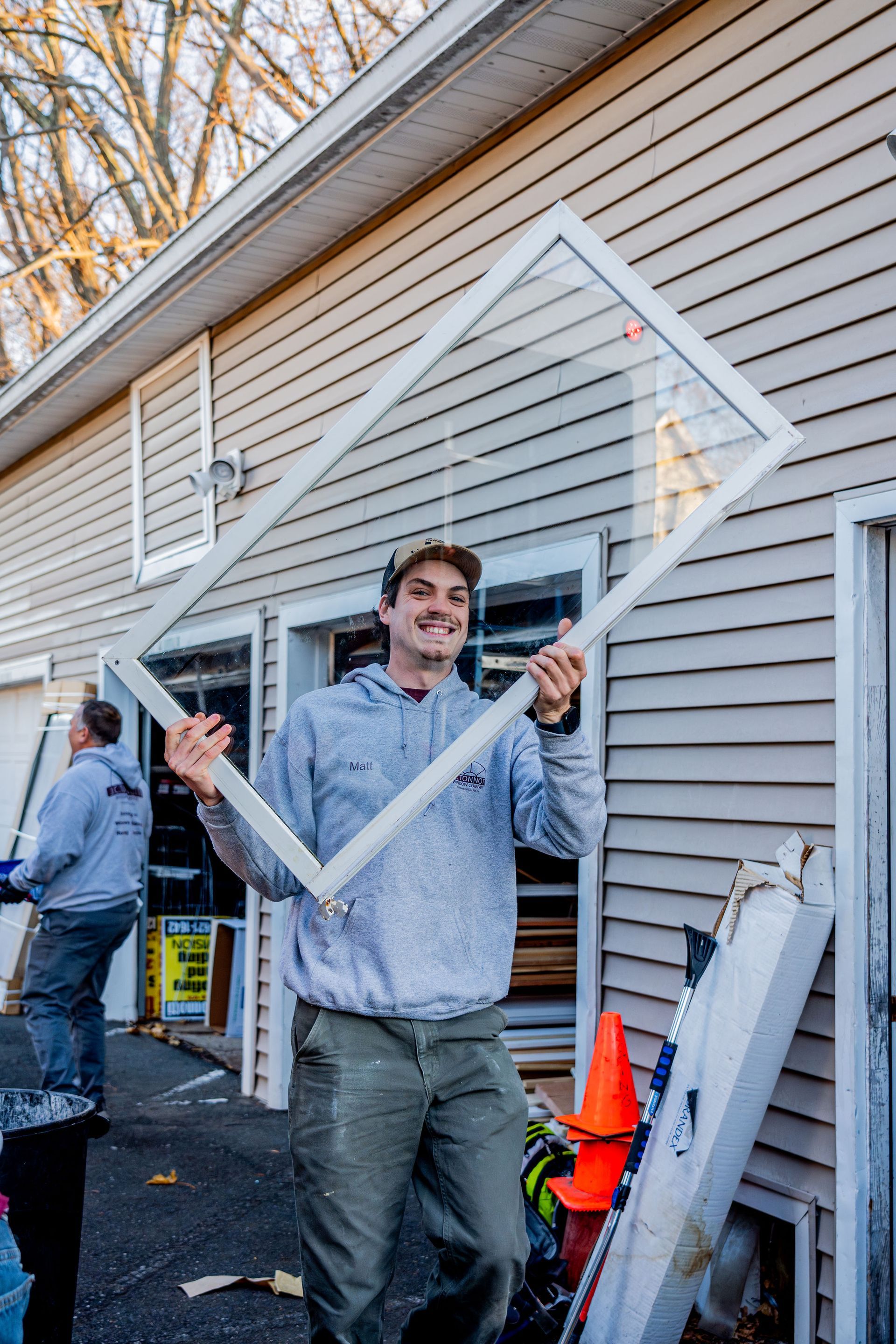 Man holding a white window frame outside a house, smiling, with another person in the background.