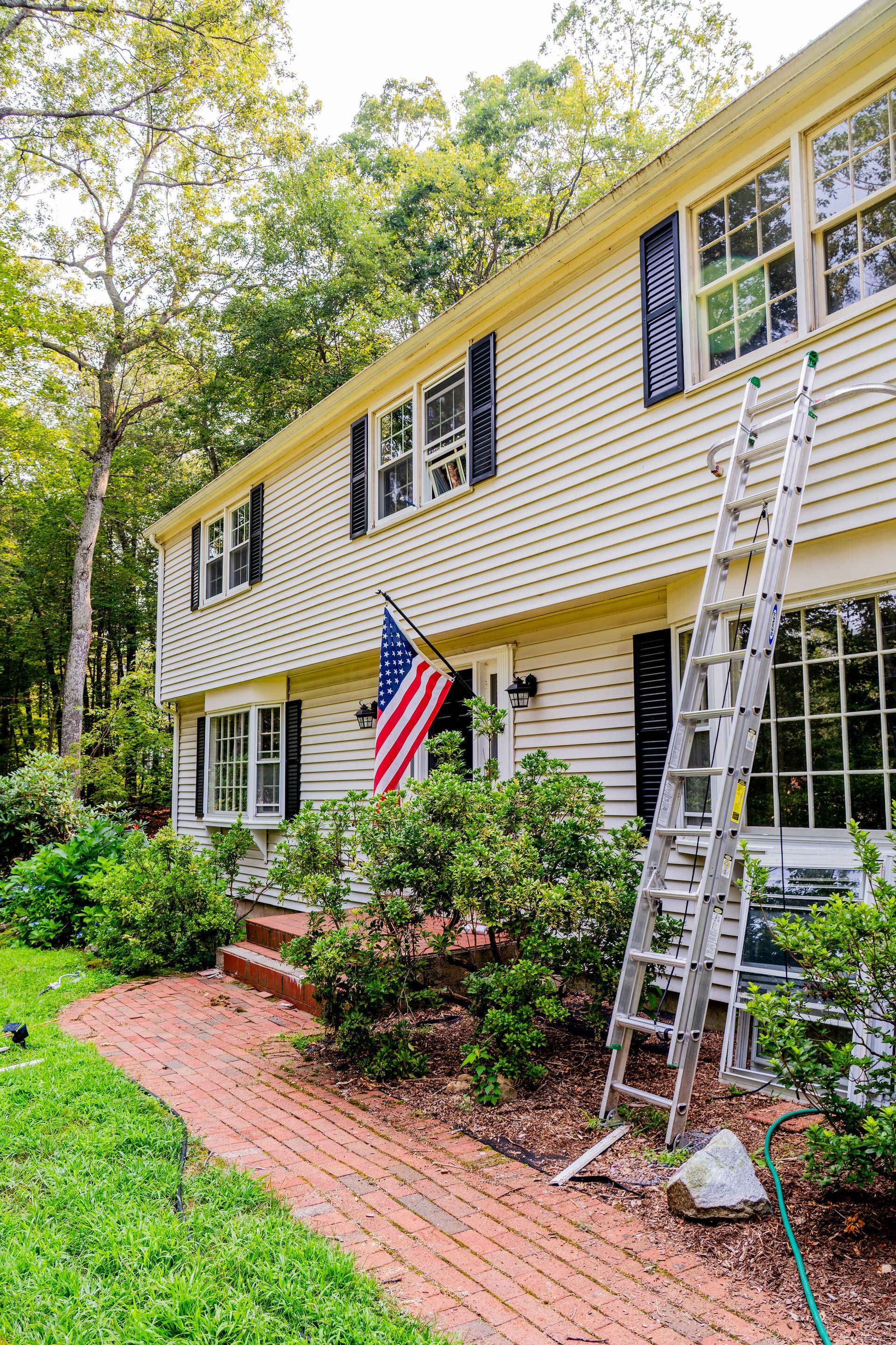 Two-story house with American flag, ladder, brick path, and greenery.