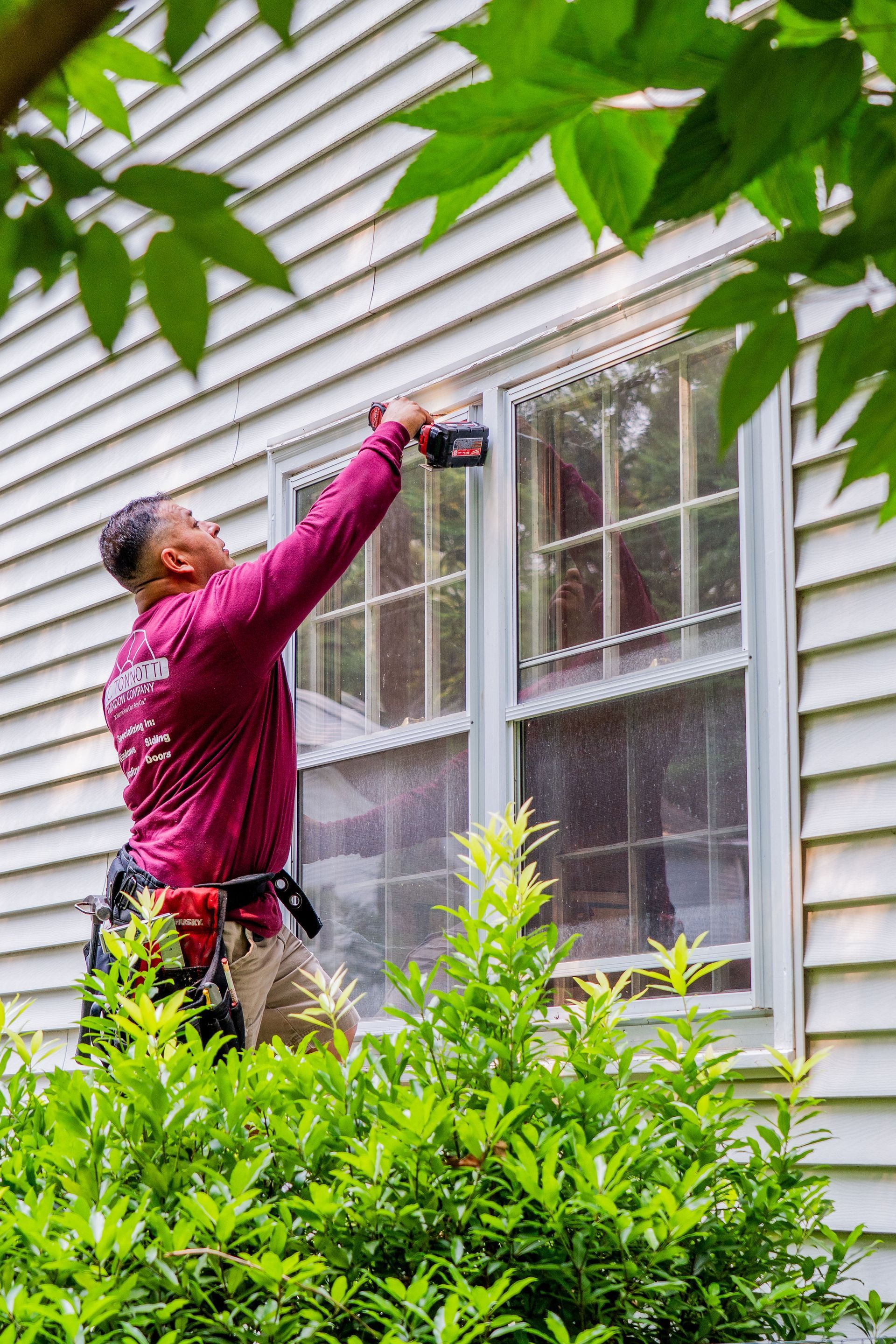 Person using a power drill on a window frame of a white clapboard house.