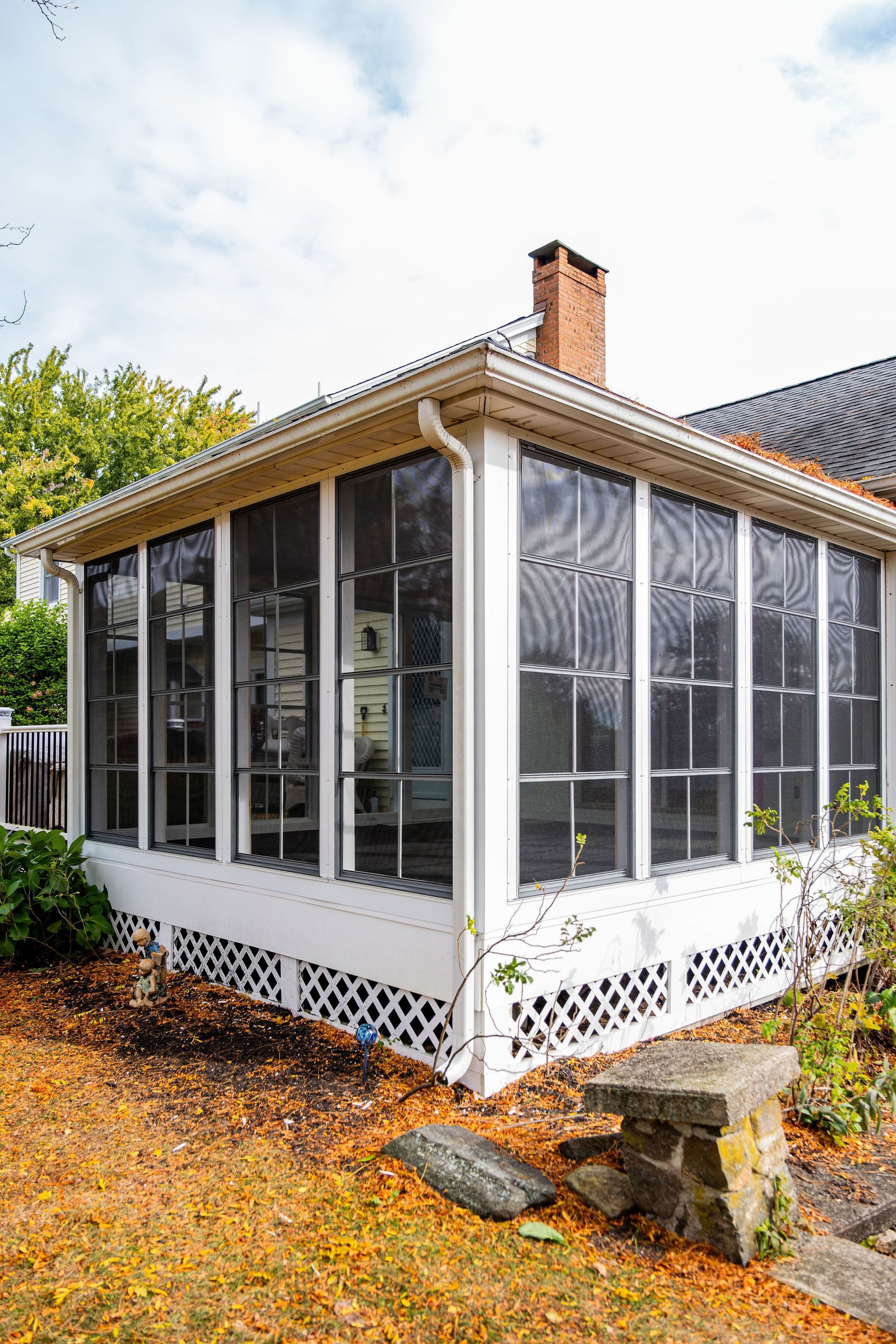 White screened-in porch with black mesh windows, chimney, and lattice detailing, set in a yard with autumn leaves.