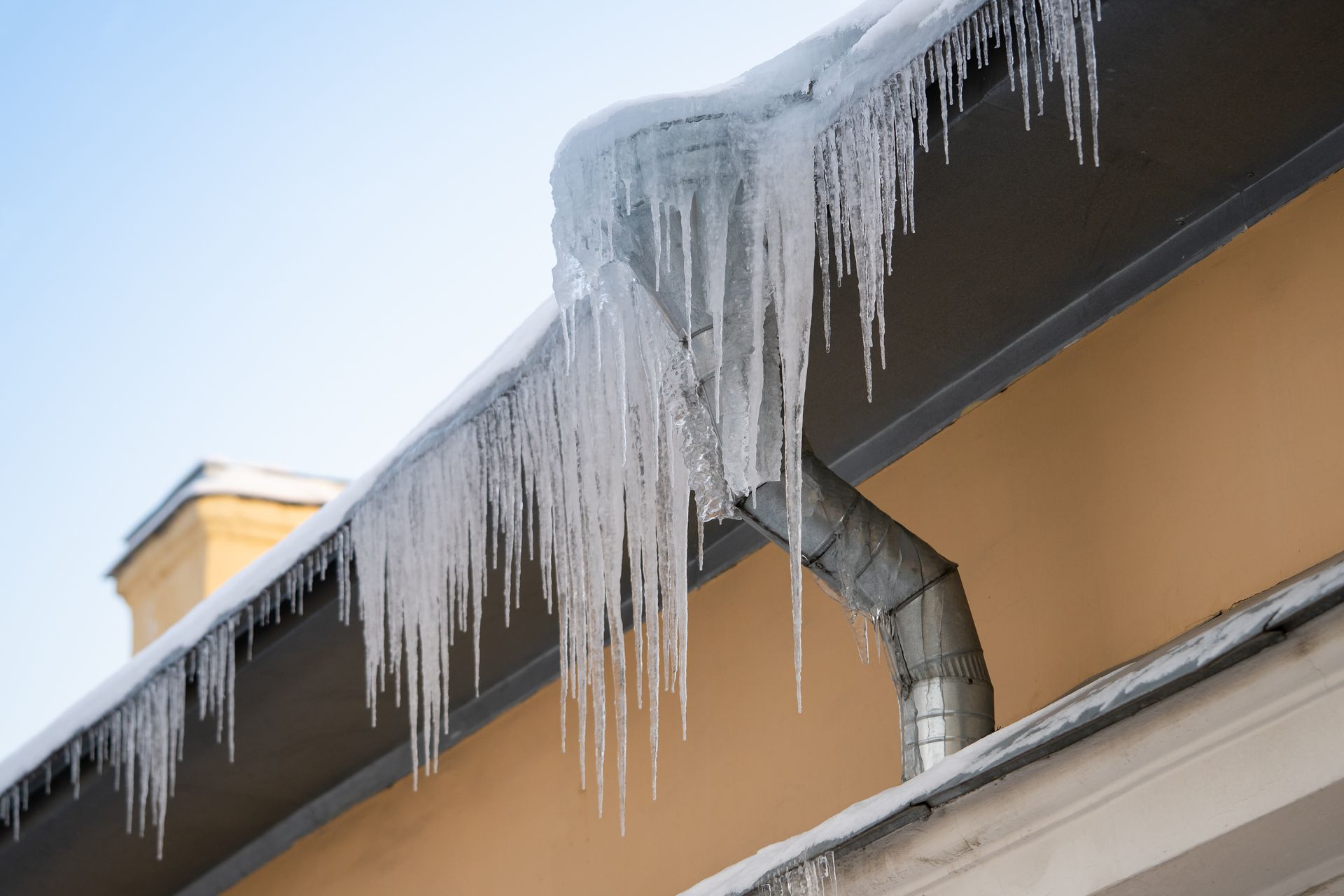 Icicles hanging from a roof gutter on a beige building against a blue sky.