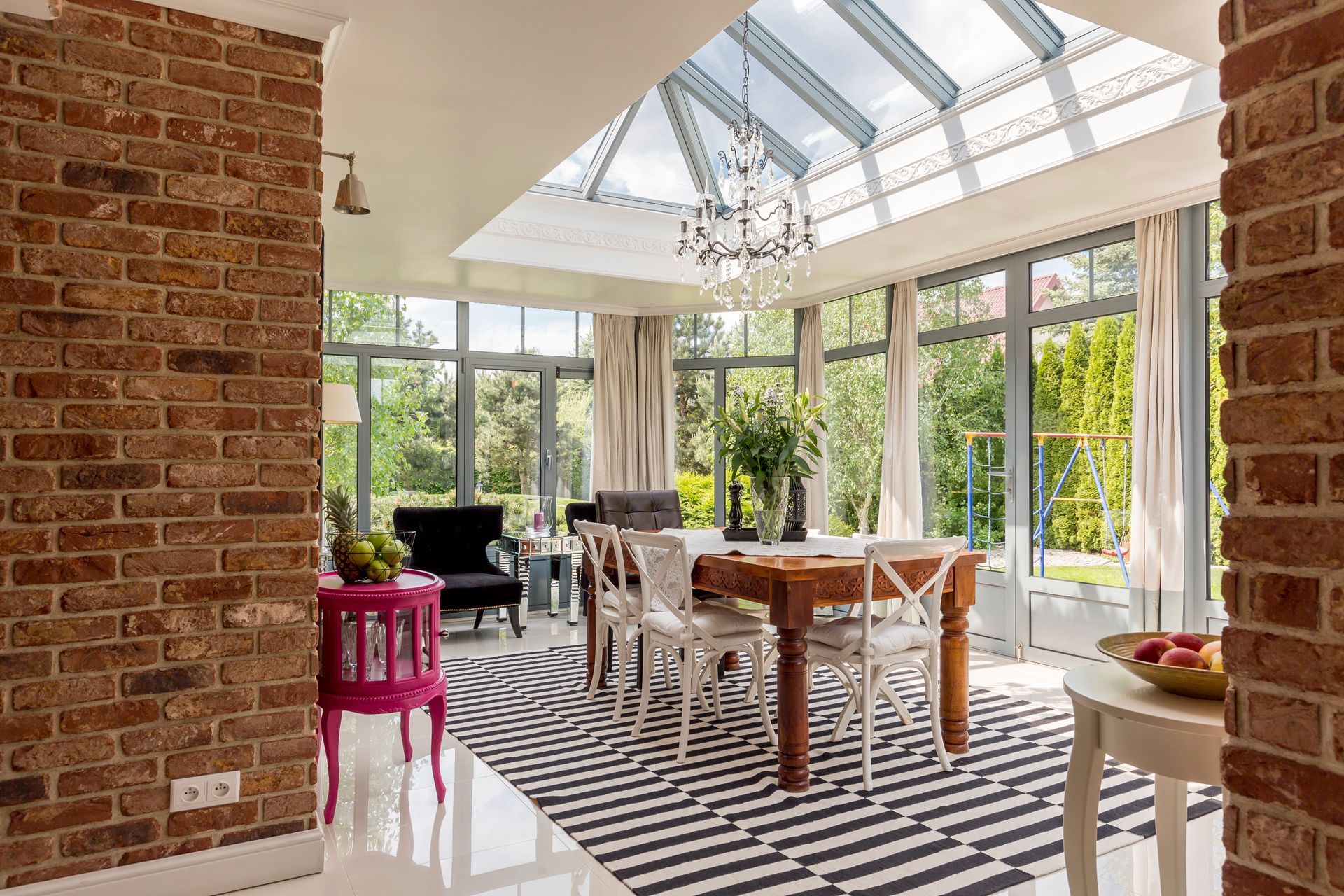 Bright sunroom dining area with a wooden table, striped rug, brick walls, and glass ceiling.