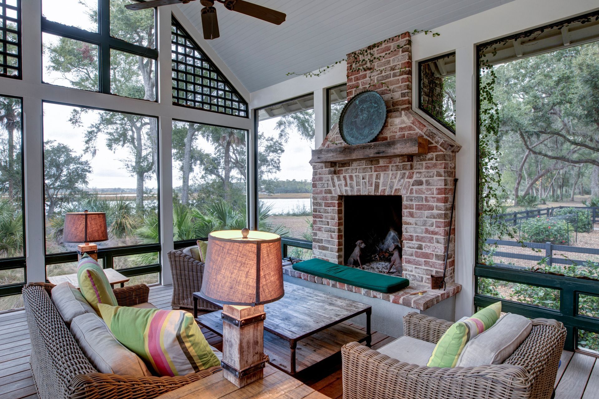 Cozy sunroom with wicker seating, a brick fireplace, large windows, and views of a wooded lakefront