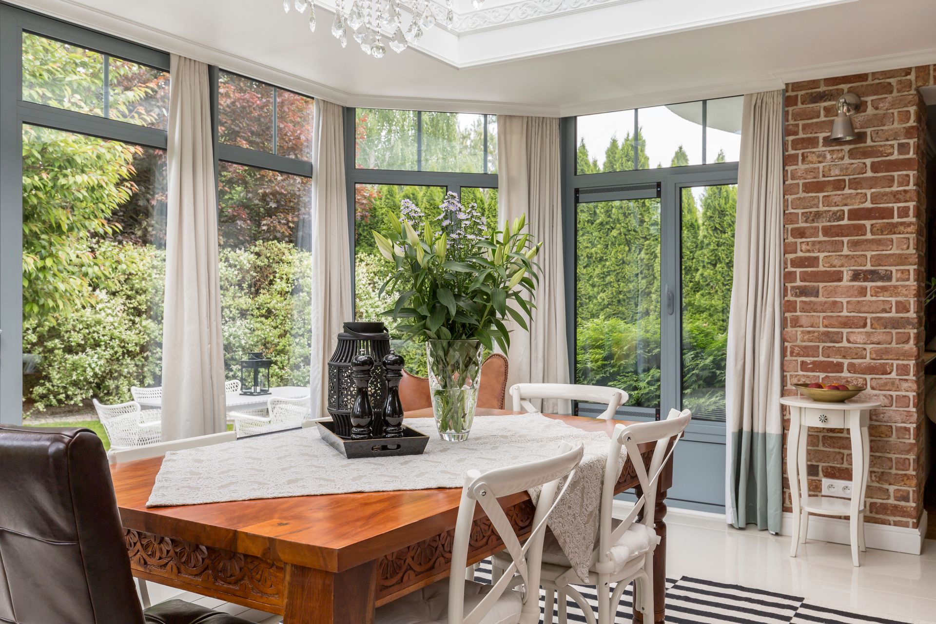 Bright dining room with large windows, white chairs, wood table, brick wall, and indoor plants.