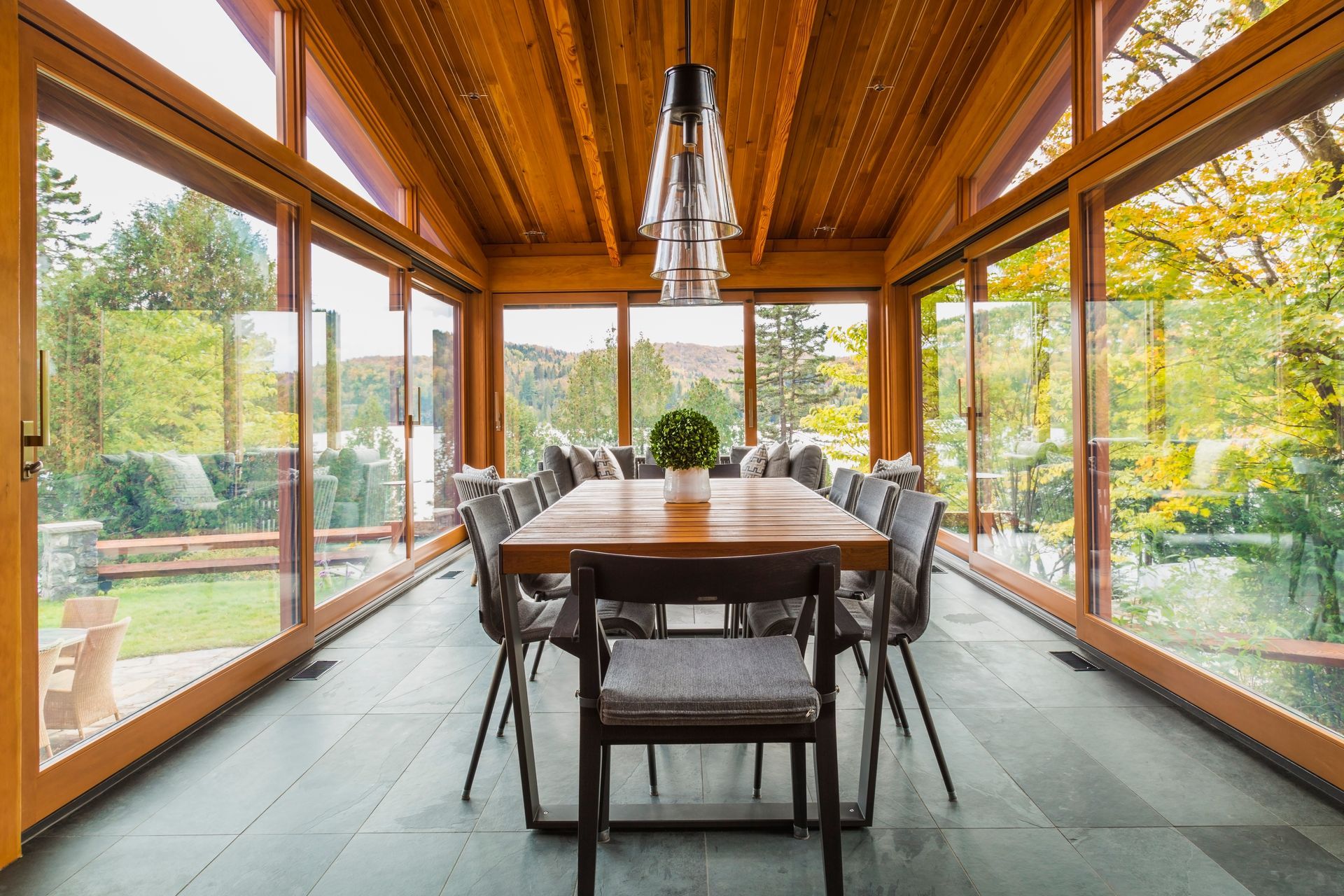Sunlit glass-enclosed dining room with a long wooden table and chairs, overlooking a green garden.