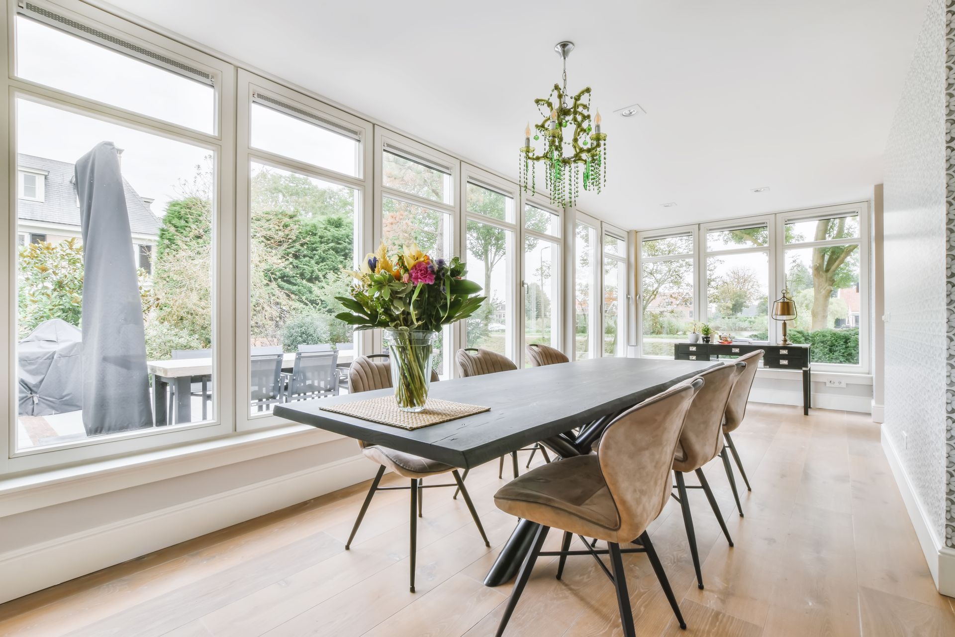 Bright dining room with large windows, wooden table, and modern chairs beside a vase of flowers