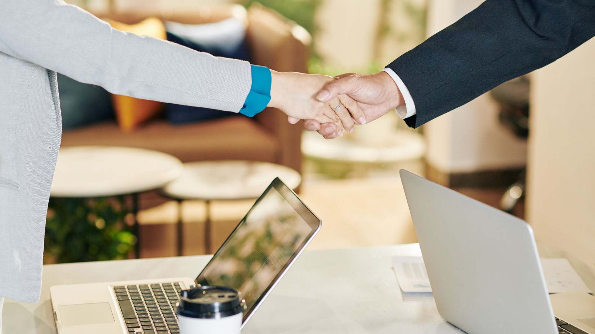 Two people shaking hands over a table with laptops, suggesting a business deal or agreement.