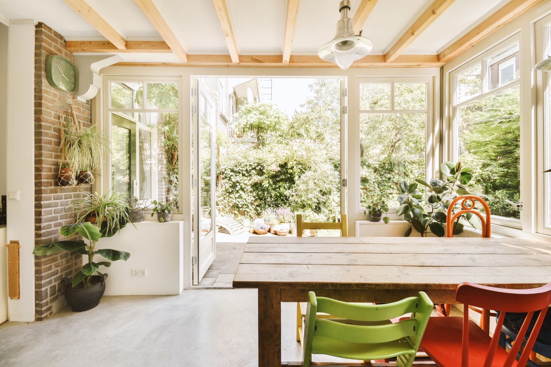 Bright sunroom with open doors, wooden table, and colorful chairs overlooking a lush garden.