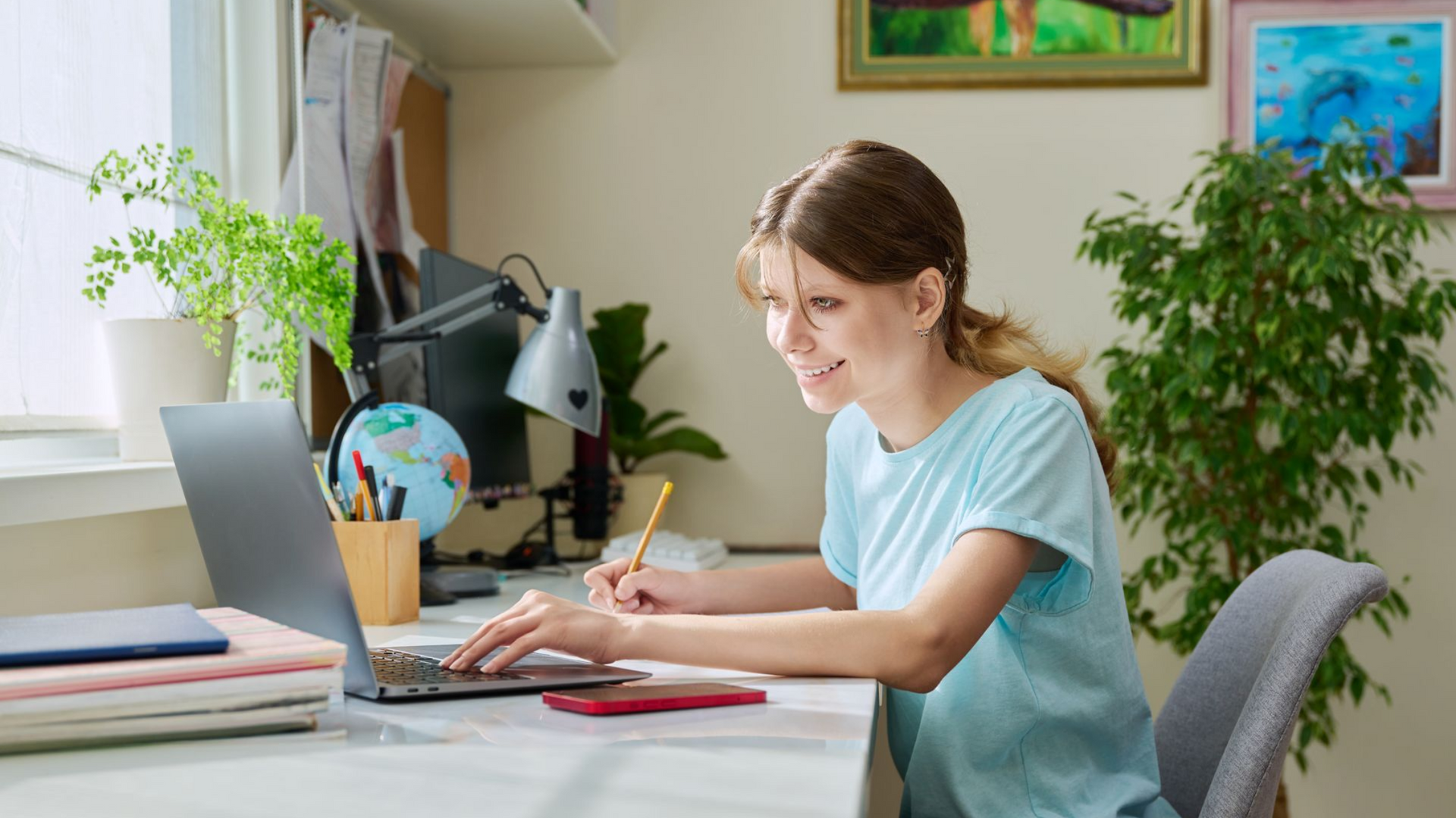 Woman smiles while working on a laptop at a desk with a globe and plants.