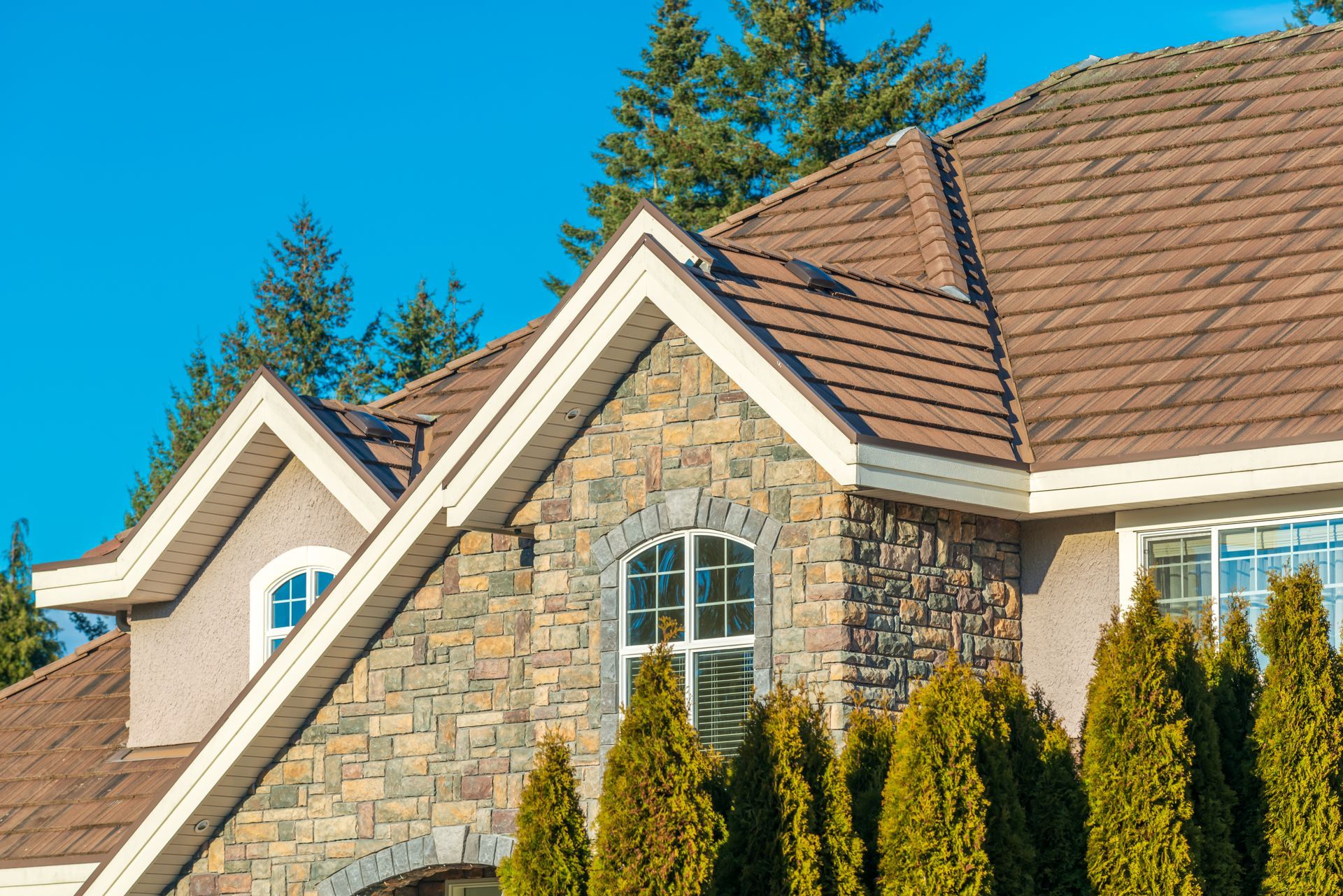 Stone house exterior with brown roof, arched windows, and tall evergreen shrubs against a blue sky