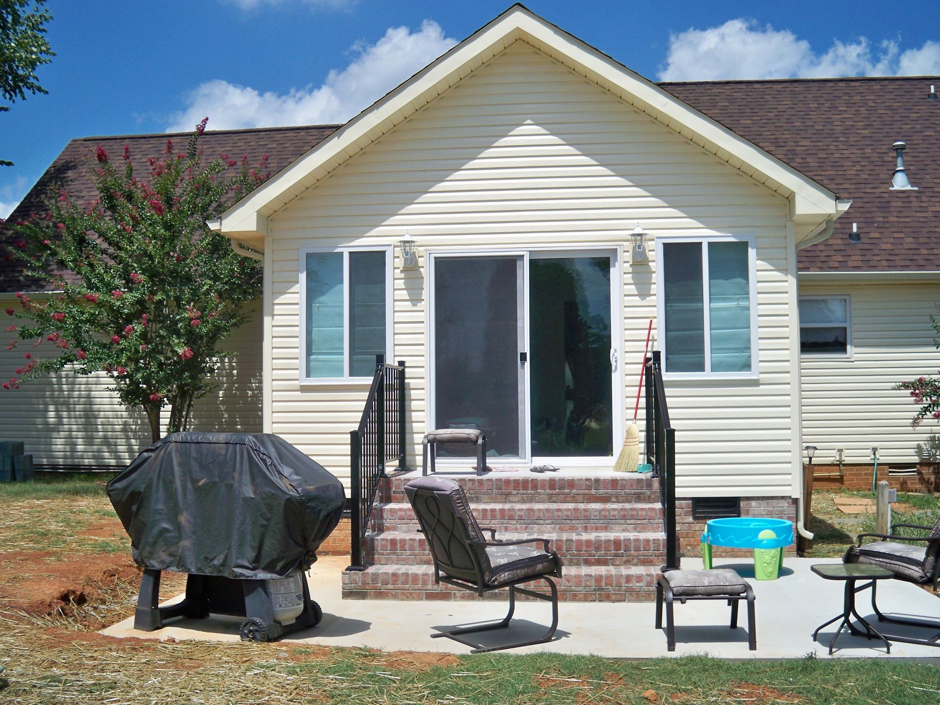Backyard patio with stairs, sliding glass door, grill, and seating. Beige siding, brown roof, and sunny outdoor setting.
