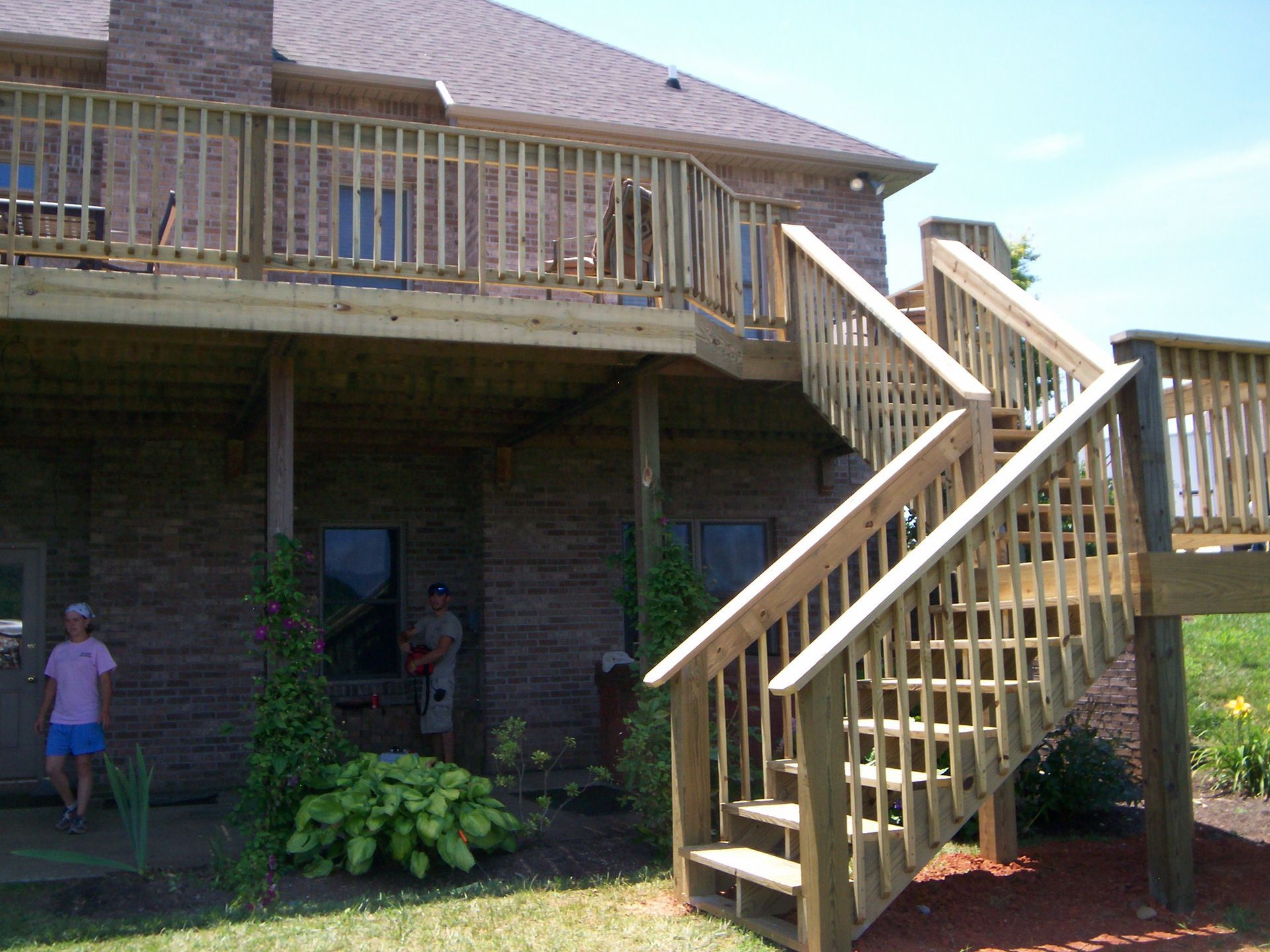 Wooden deck and stairs on a brick house. Two people visible near ground level. Sunny day.