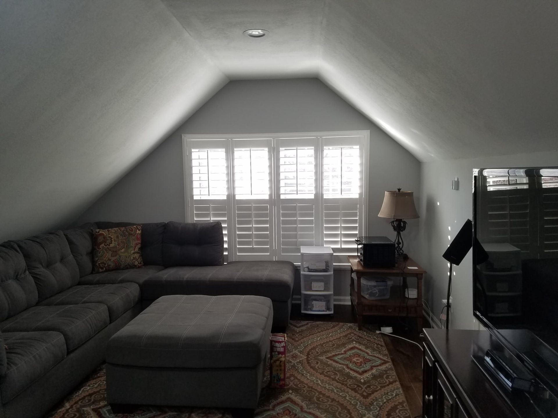 Cozy attic living room with a gray sectional sofa, ottoman, and window shutters. Dark TV and side table.