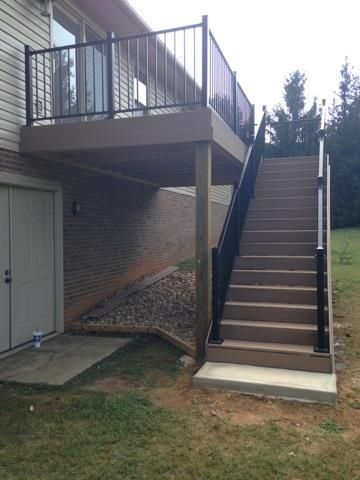 A two-level deck with stairs. The upper level has black railings. Brick wall and grass are visible.