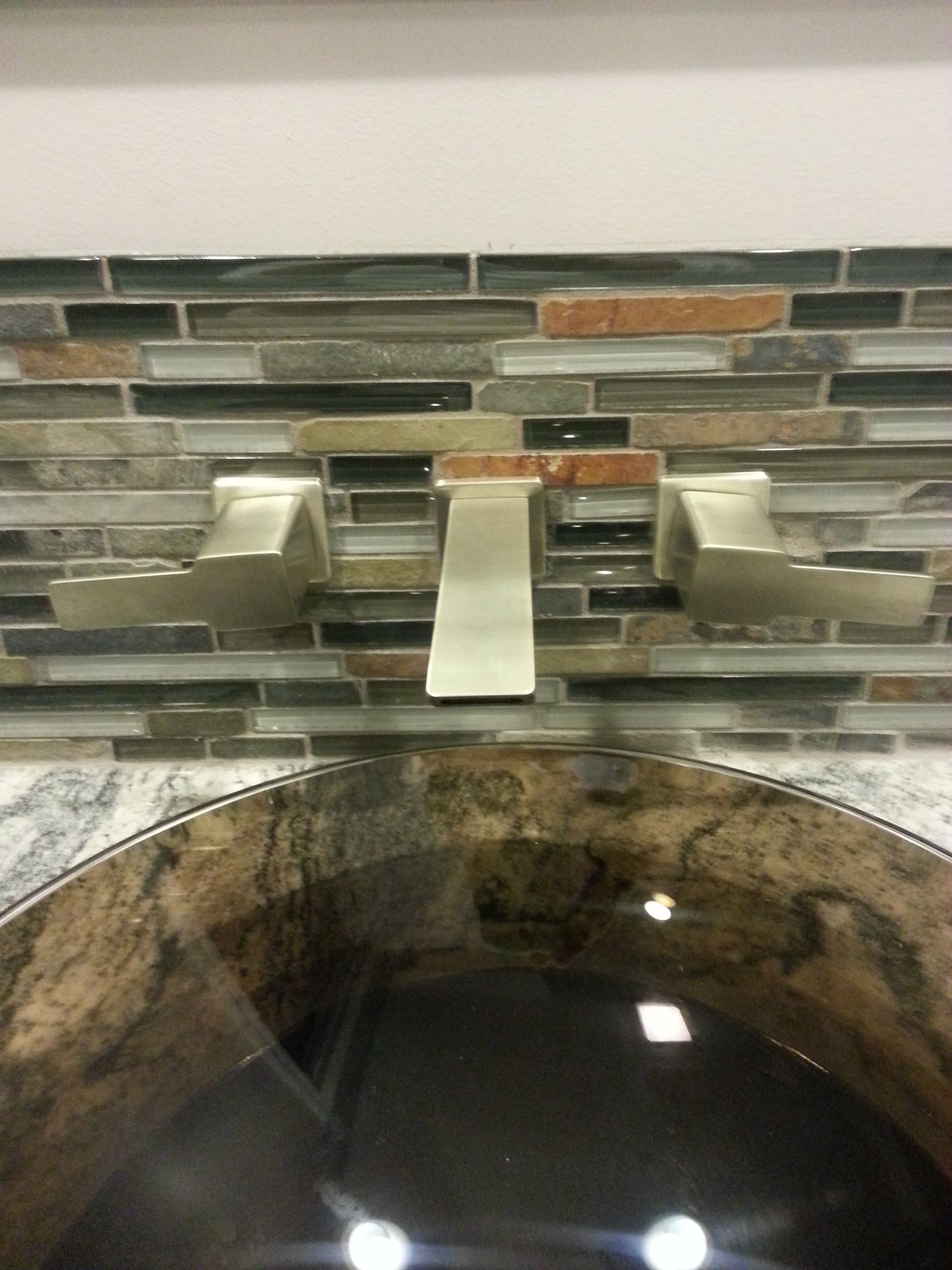 Brushed nickel faucet with lever handles over a dark, round sink, set against mosaic tile.