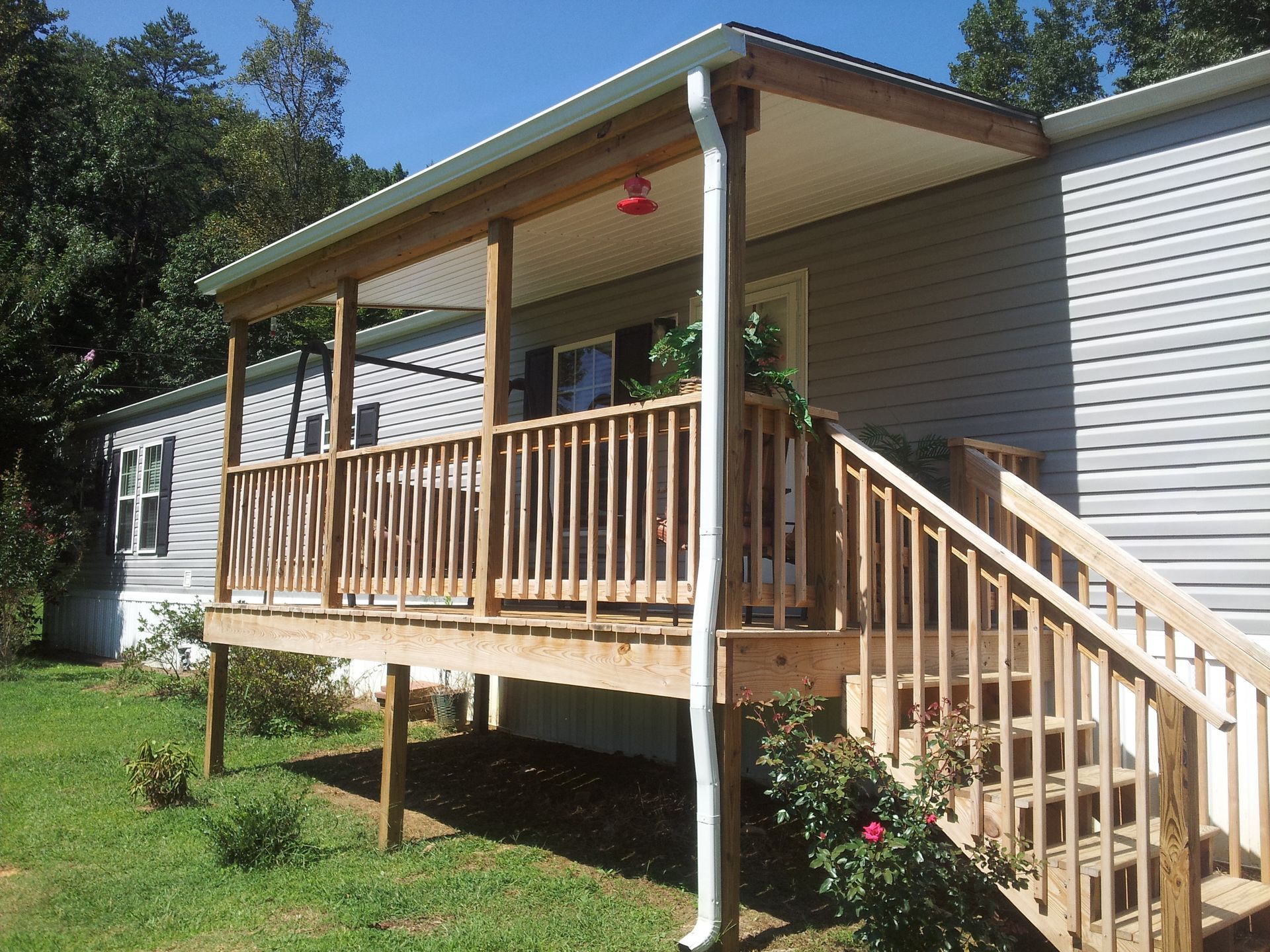 Wooden porch with a covered roof and stairs attached to a gray mobile home on a sunny day.