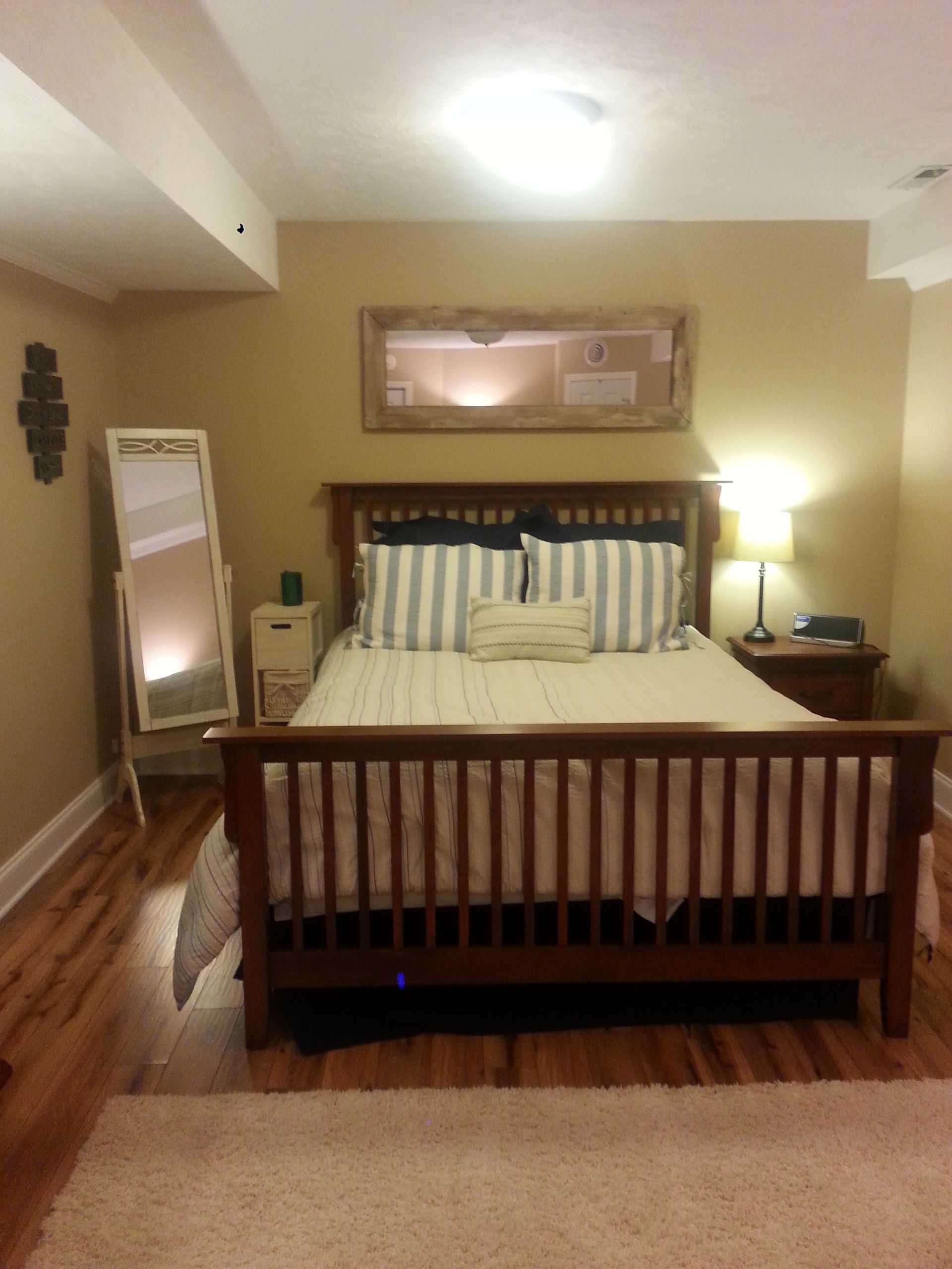 Bedroom with wooden bed, full-length mirror, nightstand, and rug on a hardwood floor.