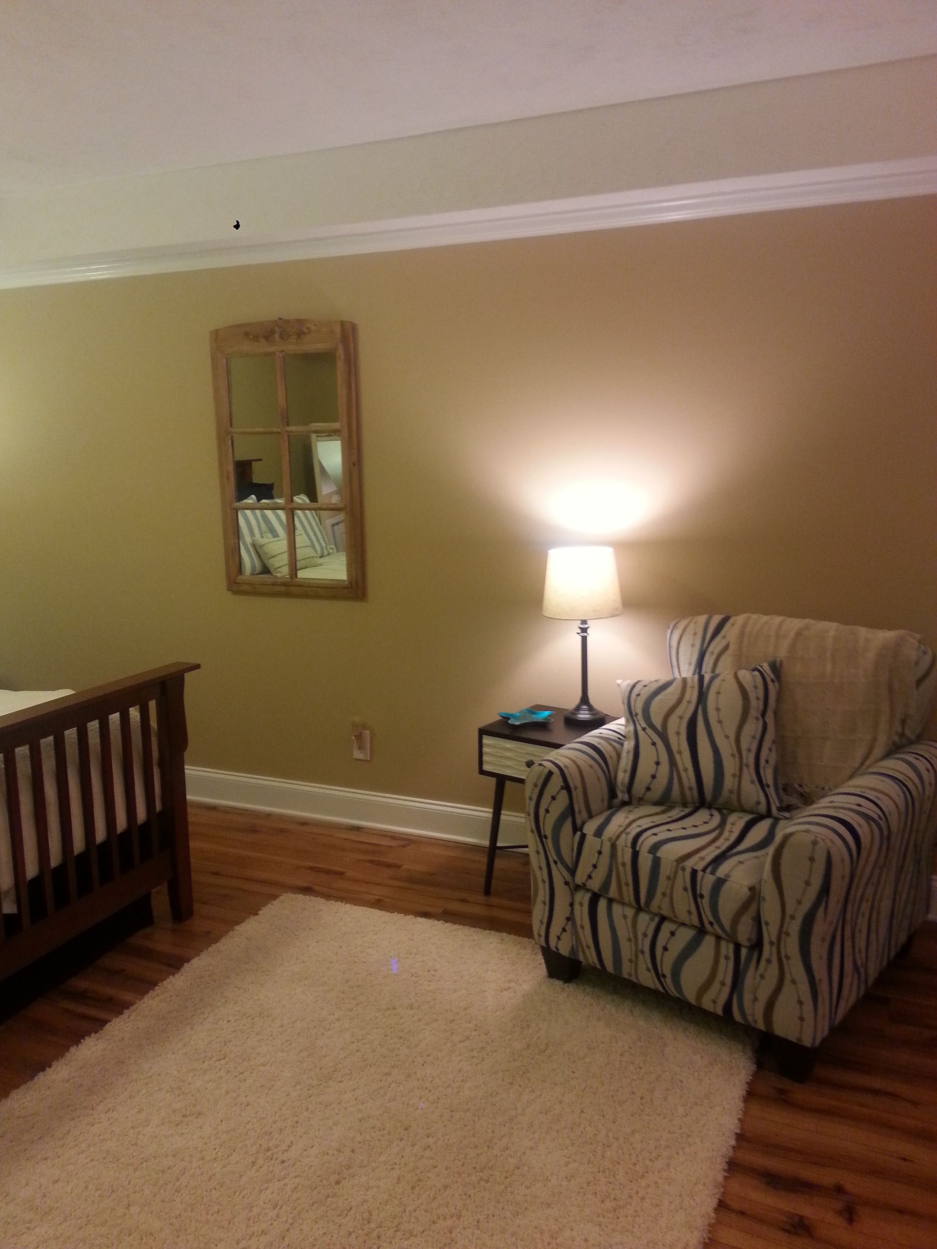A bedroom with a patterned armchair, bedside table, rug, mirror, and crib. Brown walls and hardwood floors.