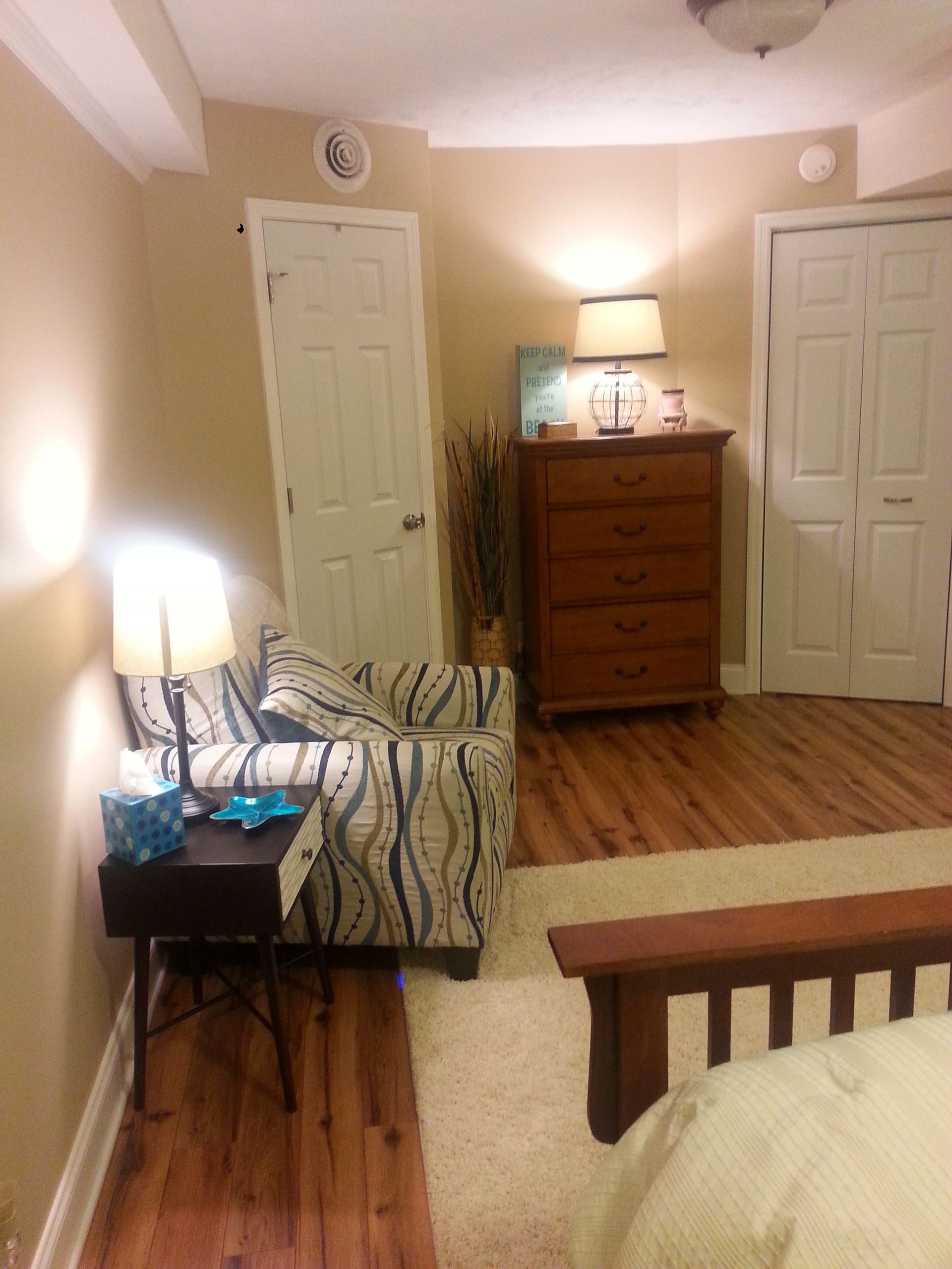 Cozy bedroom corner with armchair, lamp, nightstand, and chest of drawers. Wooden floor, beige walls, and white doors.