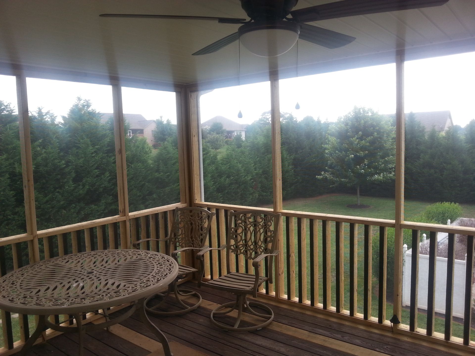 Screened porch overlooking a backyard with trees, table, chair, and ceiling fan.