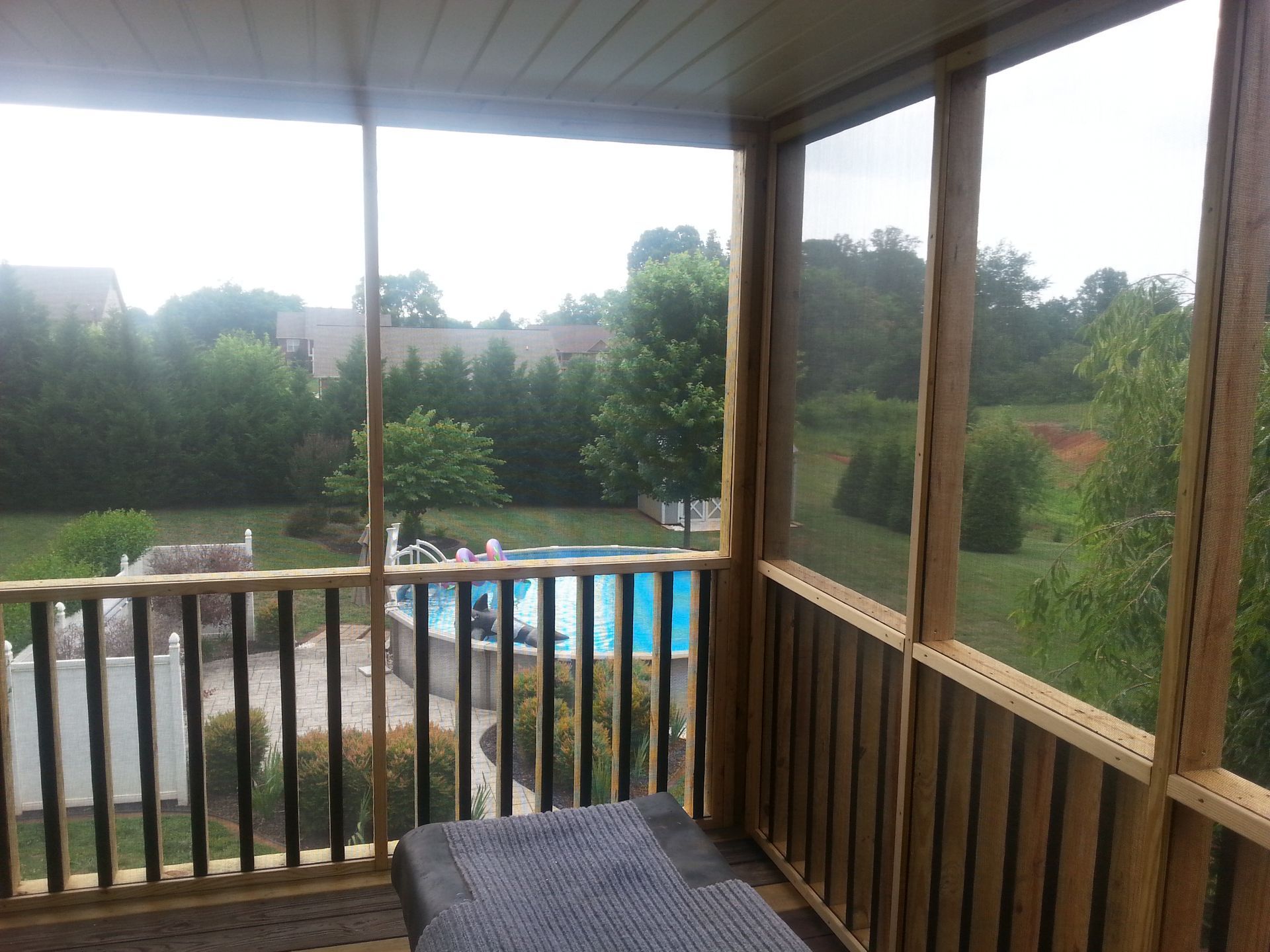 Screened-in porch overlooking a pool and yard with trees. Brown wood, blue water, and green foliage visible.
