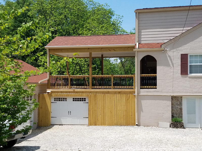 Two-story house with a wooden deck above a garage; light beige walls, red roof, and gravel driveway.
