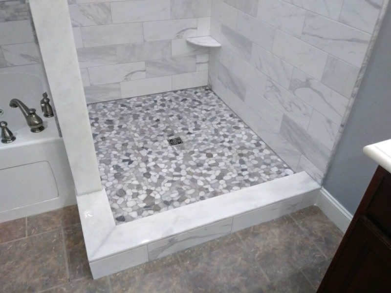Shower stall with pebble tile floor and marble walls, next to a bathtub.