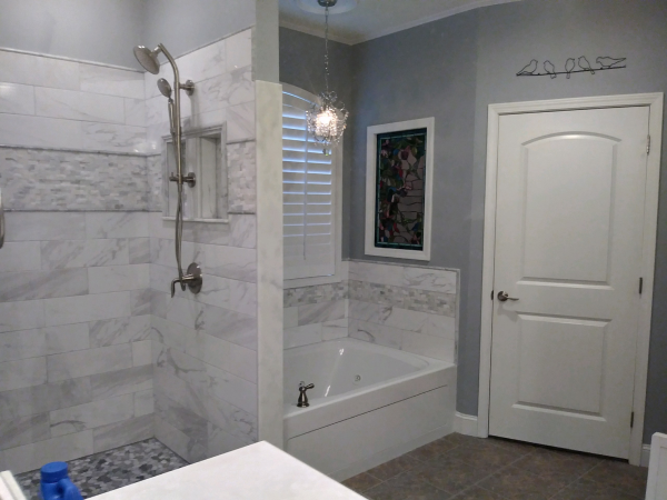 Bathroom with a tiled shower, white tub, and grey walls. A window is in the center, and a white door is to the right.