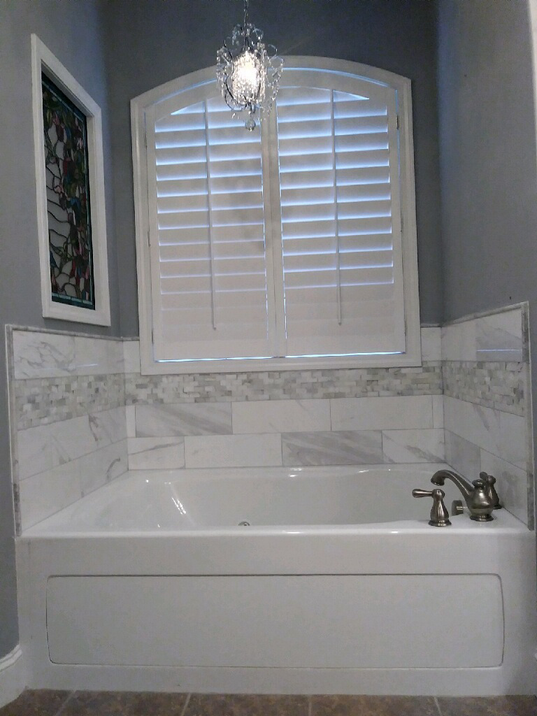 White bathtub under a window with white shutters, gray walls, and a decorative light fixture.