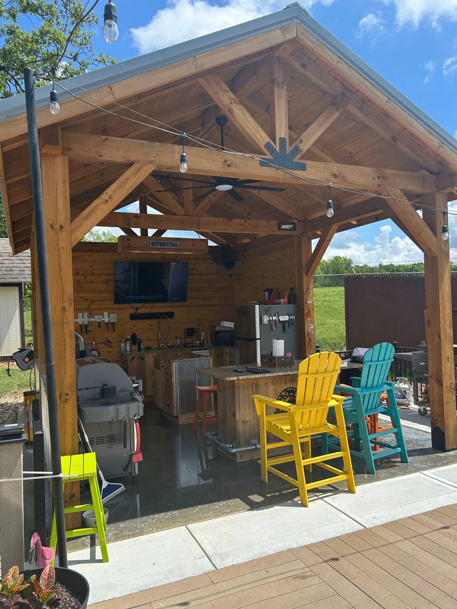 Wooden outdoor kitchen with a bar, seating, appliances, and a TV; chairs are yellow and teal.