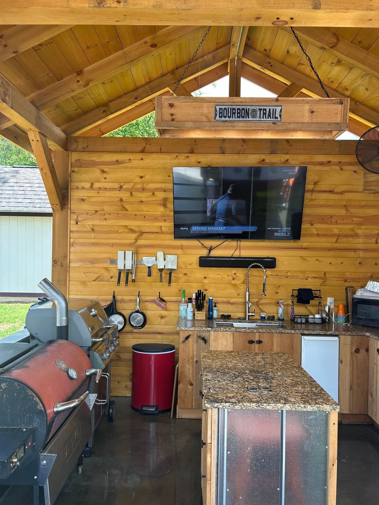 Outdoor kitchen with a smoker, grill, TV, and kitchen island under a wood-beamed pavilion.