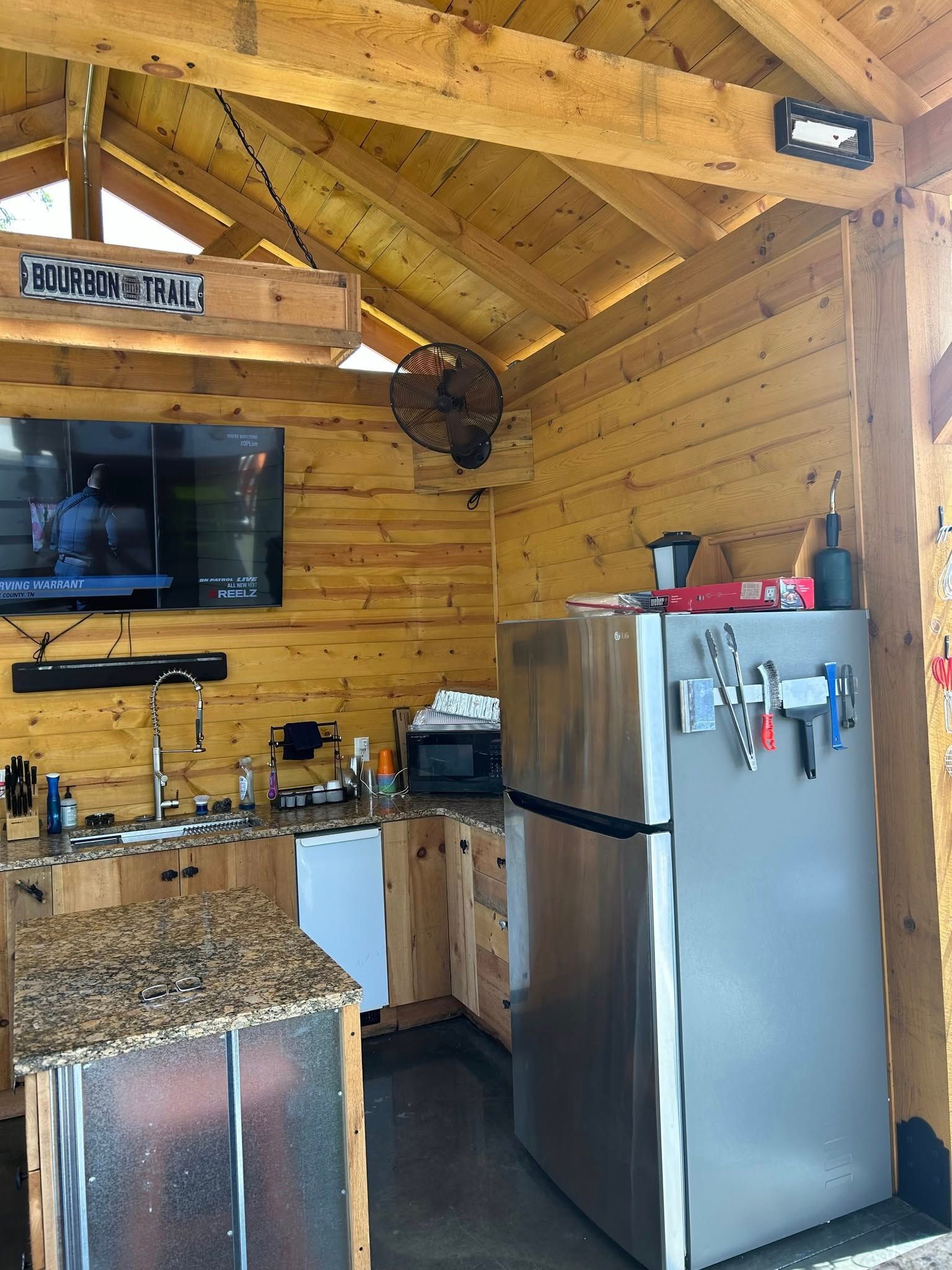Outdoor kitchen with wood paneling, granite countertops, and stainless steel appliances. A television and fan are mounted.