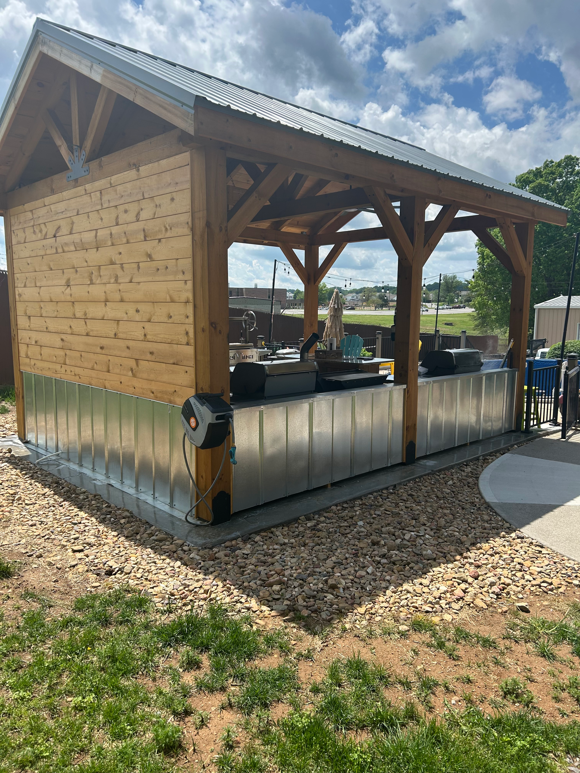 Wooden outdoor kitchen with metal countertops and roof under a cloudy sky.