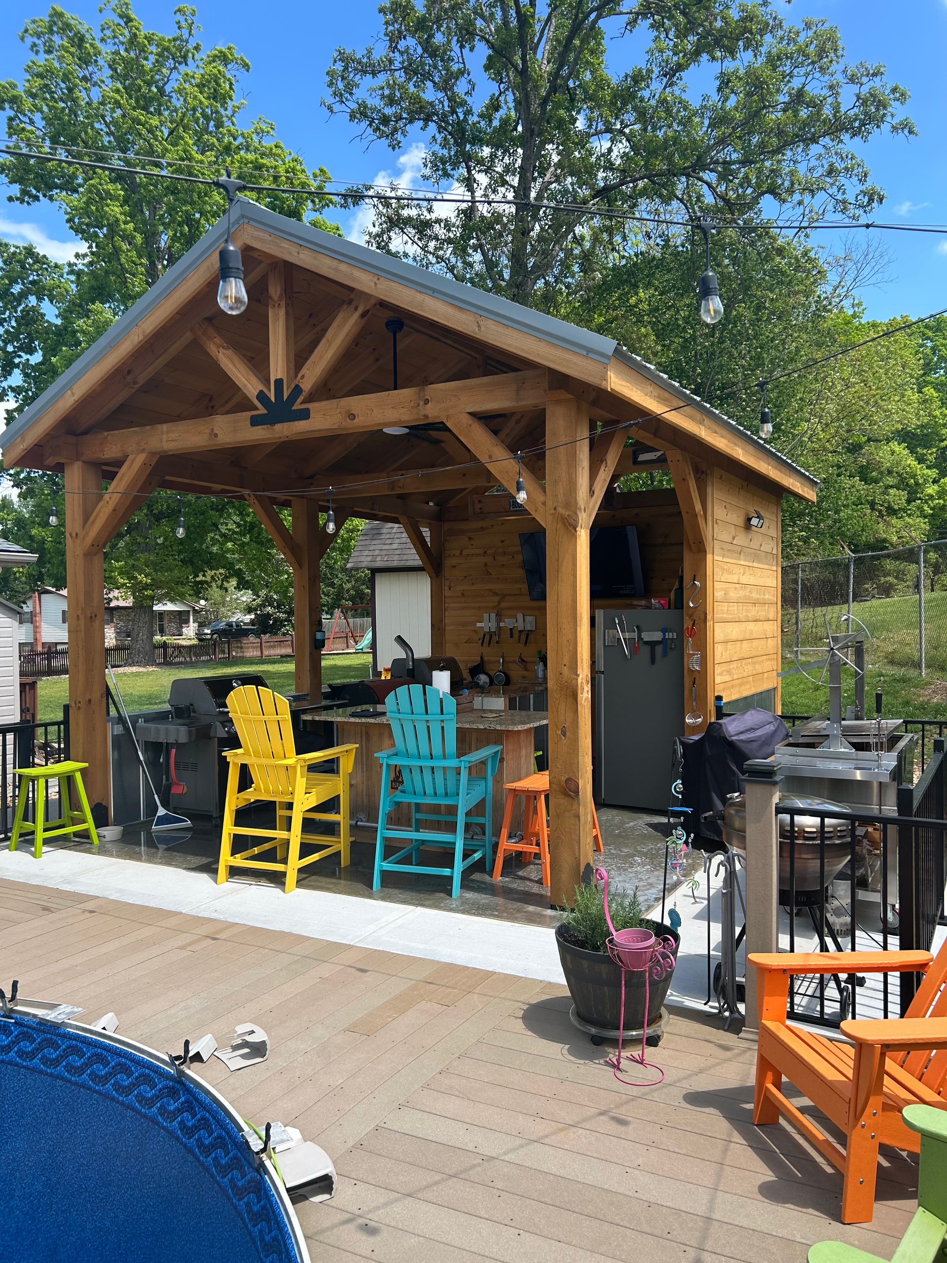Outdoor kitchen with wooden structure, colorful chairs, grill, and appliances.