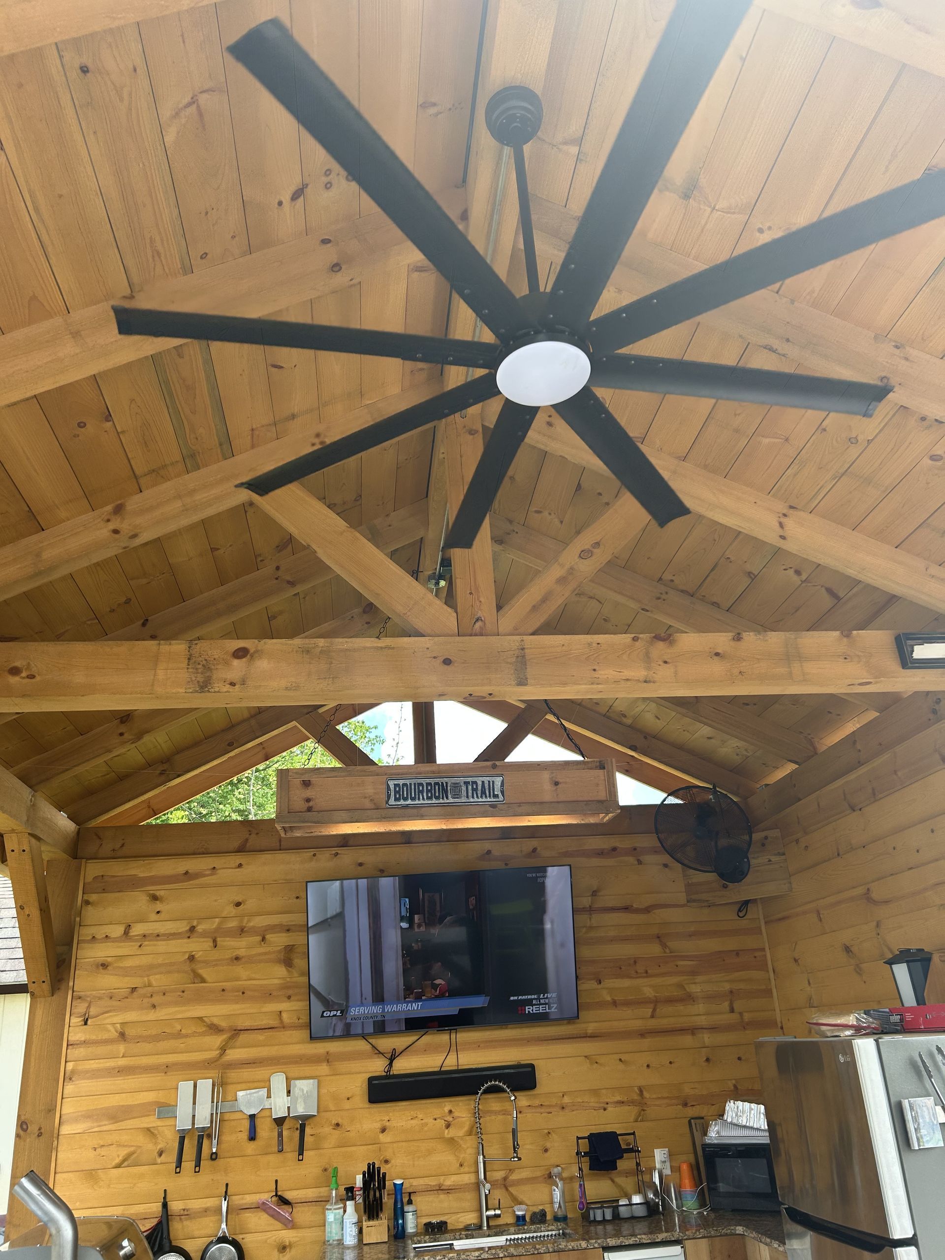 A wooden outdoor structure with a large black ceiling fan and a TV mounted on a wooden wall.