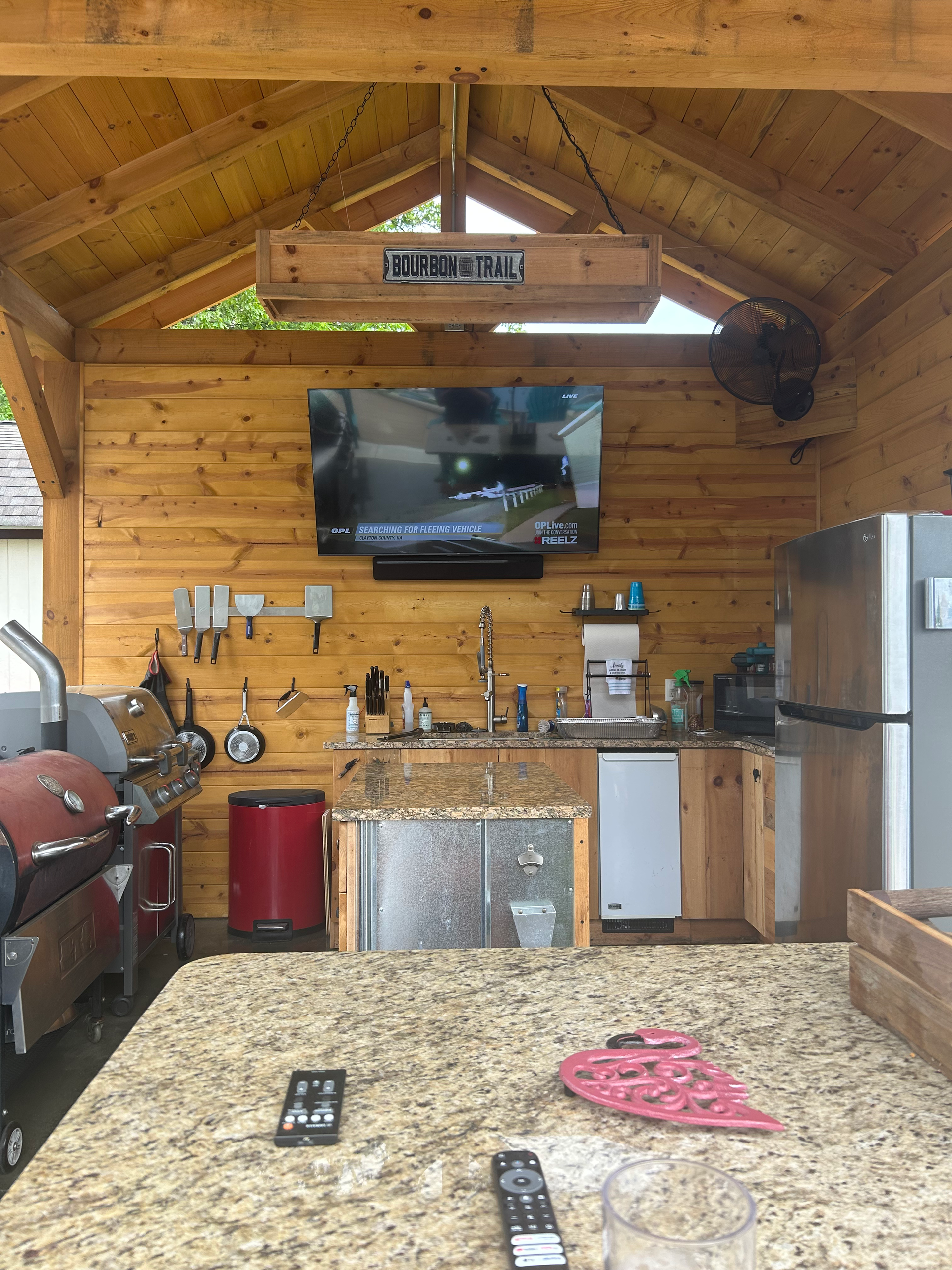 Outdoor kitchen with a TV, grill, and countertop. Wooden walls and roof.